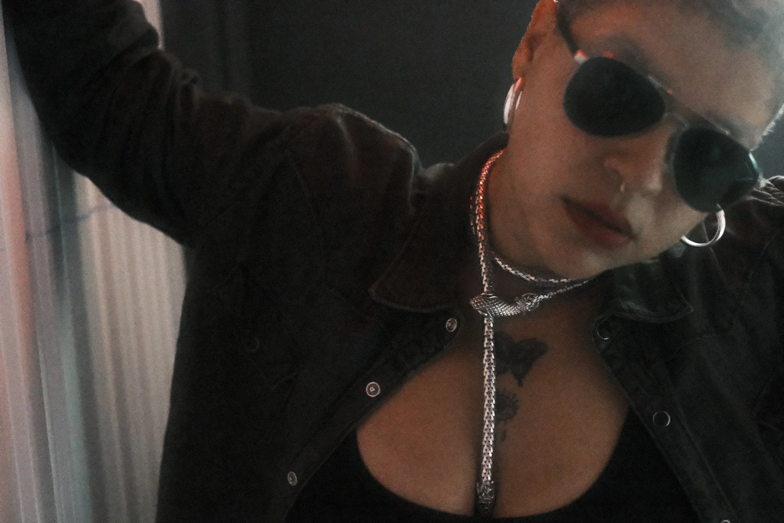 A person wearing sunglasses, a chain necklace, earrings, and a dark jacket, with makeup including lipstick, appears to be in a dimly lit environment.