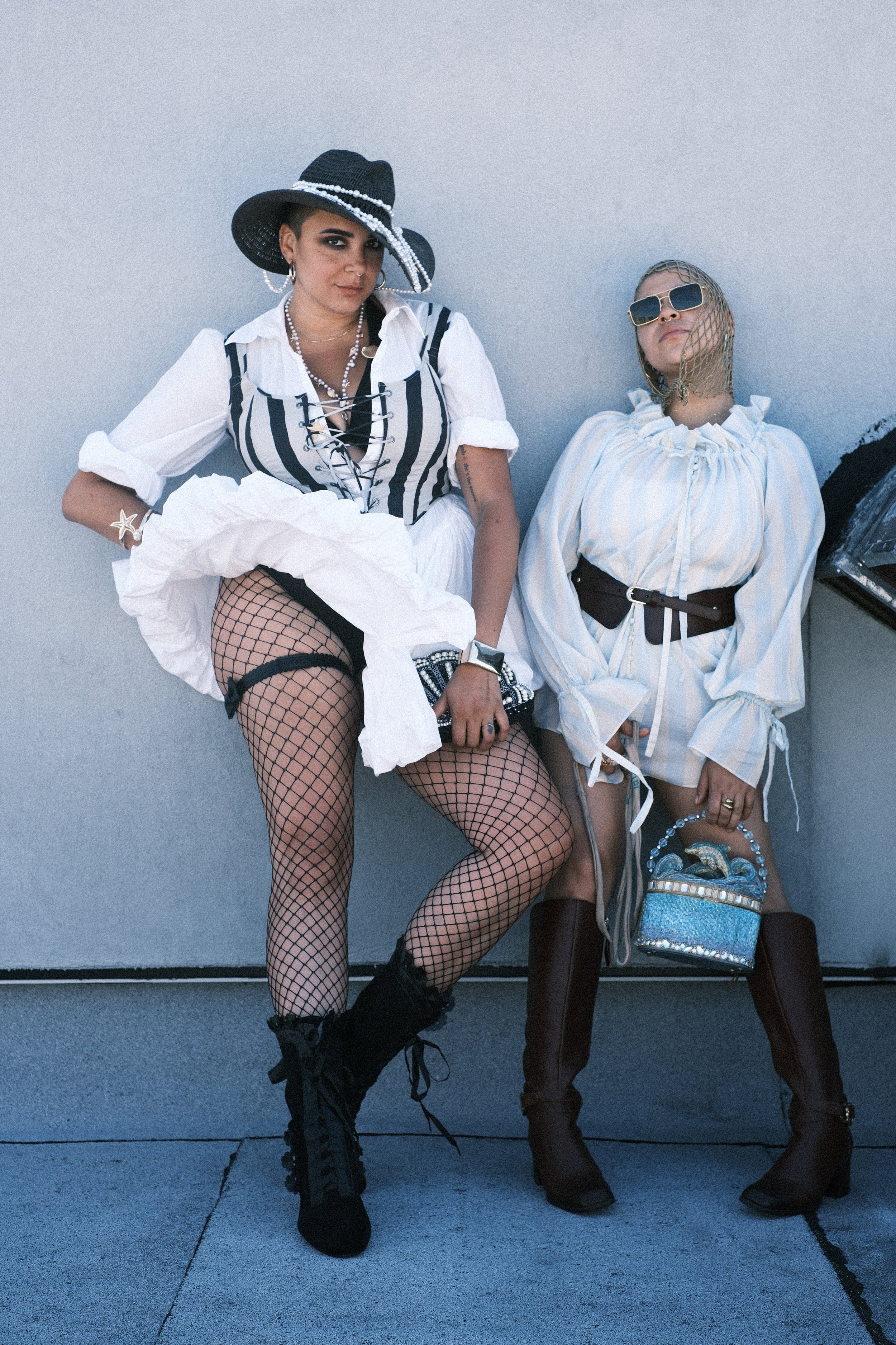 Two women are sitting against a plain blue wall. The woman on the left is wearing a black wide-brimmed hat with pearls, fishnet stockings, black boots, and a striped black and white shirt with lace-up detail. The woman on the right has short blonde h
