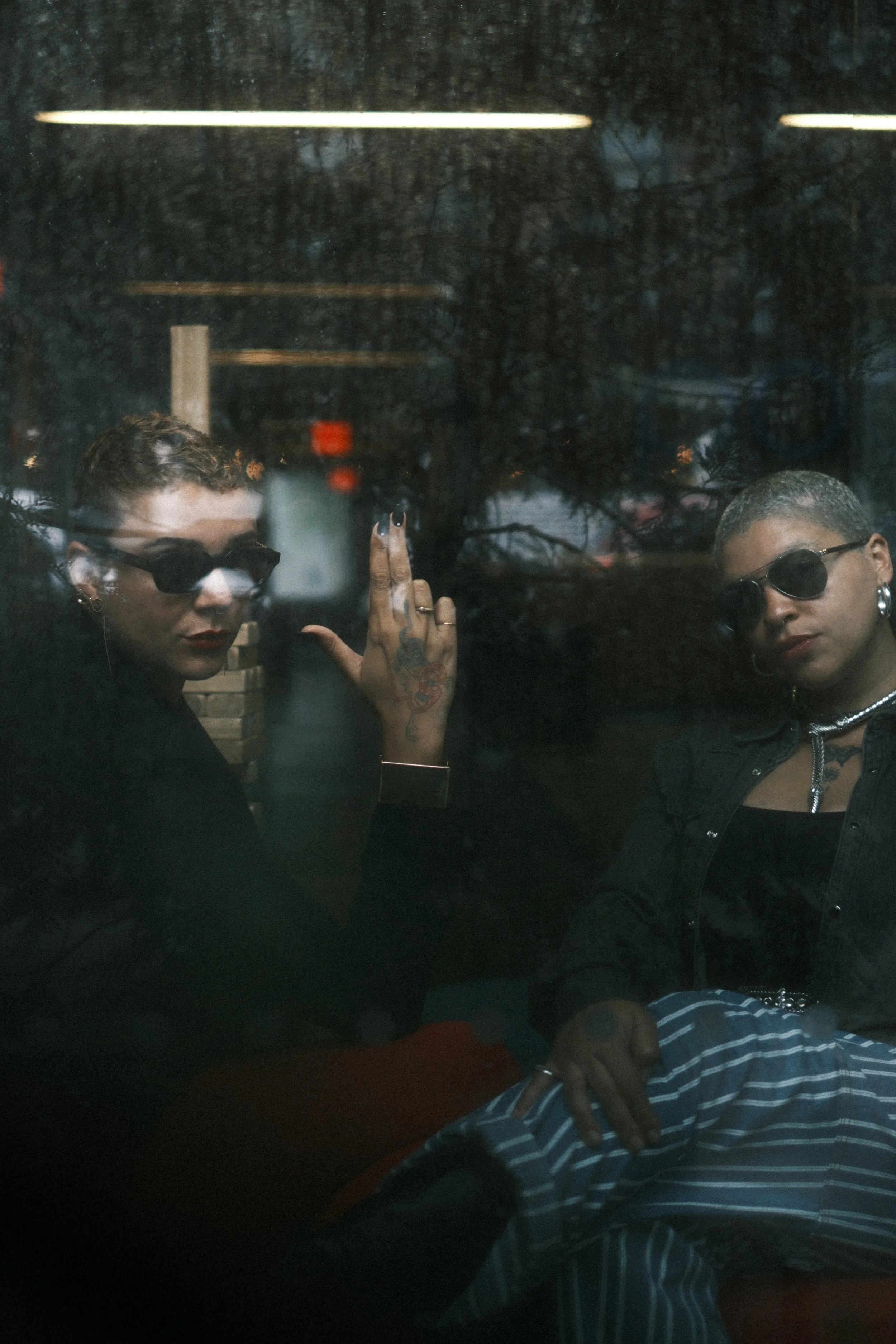 Two women with short hair and sunglasses are sitting behind a reflective window, possibly in a cafe or similar setting. One woman has tattoos on her hand and is wearing earrings, while the other is dressed in a striped shirt.