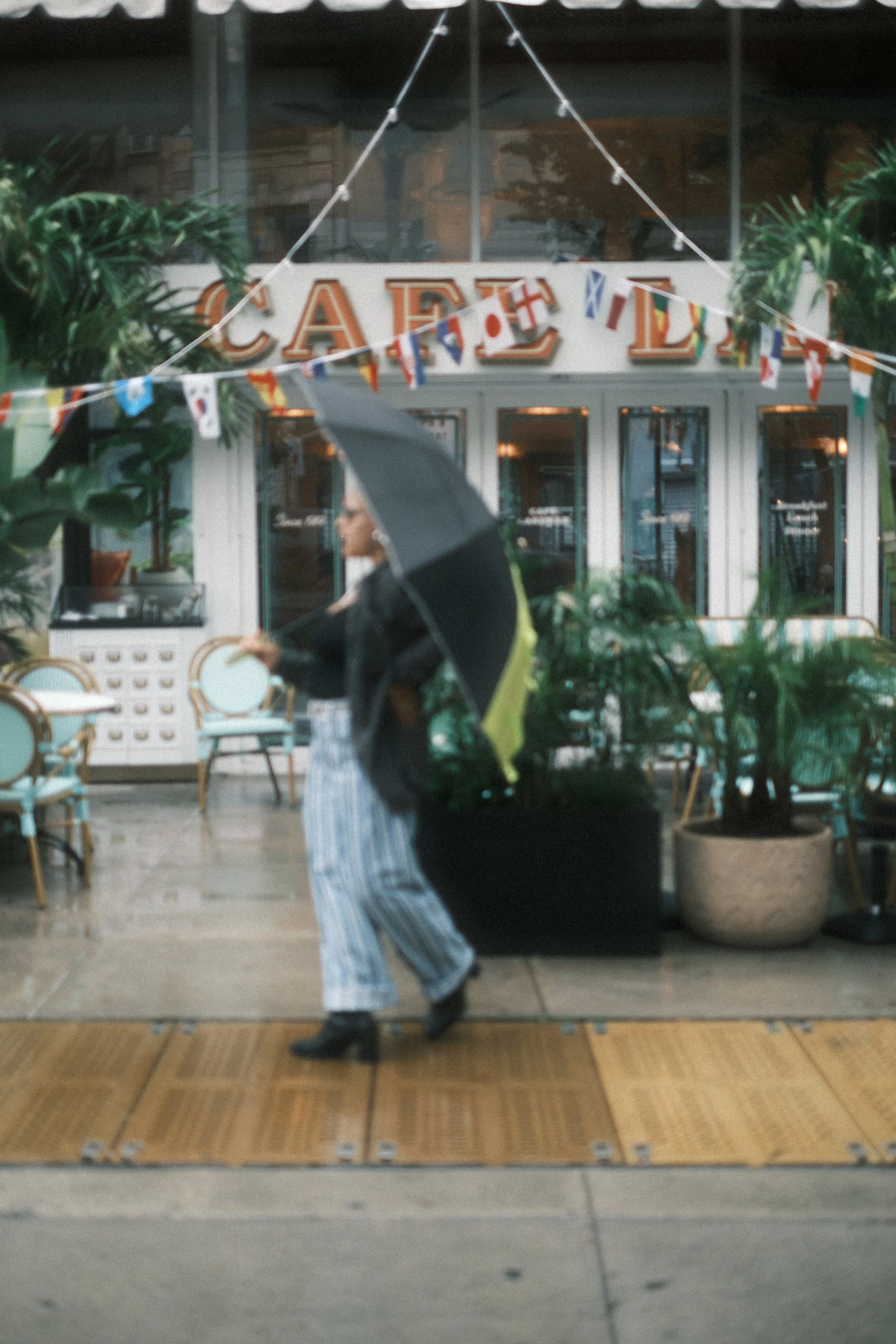 Person walking with an umbrella in front of a cafe, with outdoor seating and plants, on a rainy day.