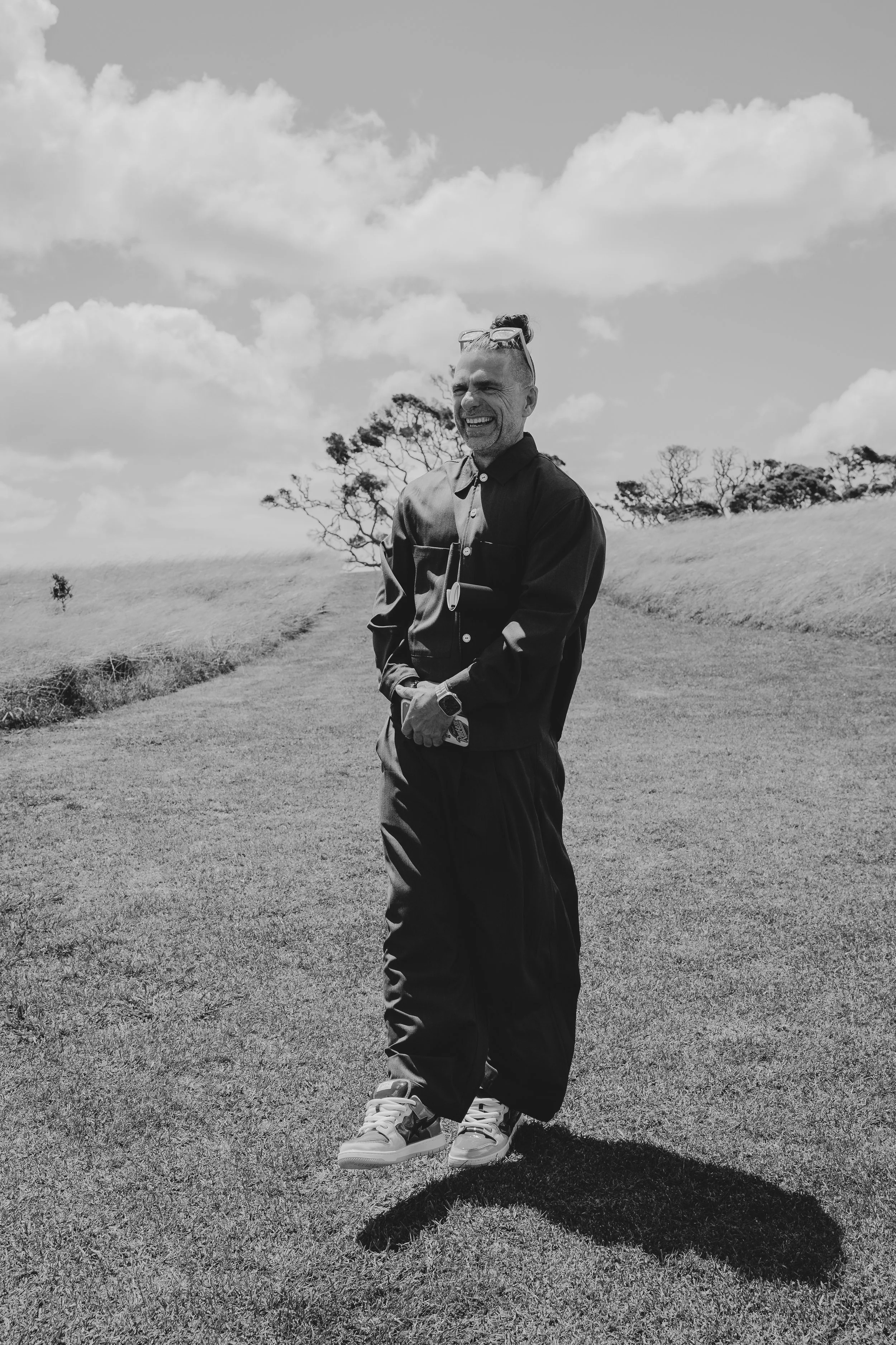 A smiling man with sunglasses on his head, wearing a dark hoodie and pants, standing on a grassy hill with trees in the background and a partly cloudy sky above.