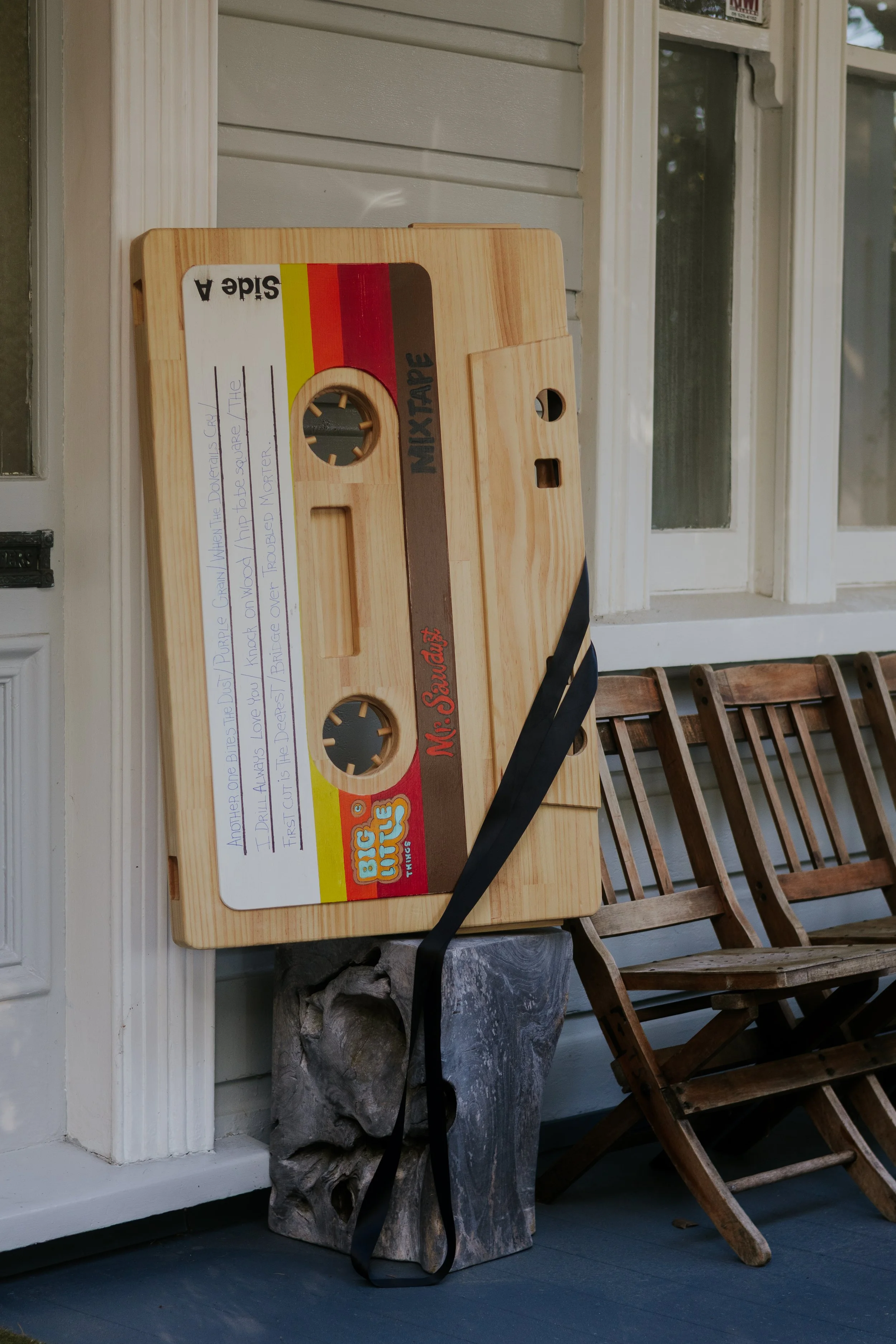A wooden cassette tape storage box placed on a rock, with a handwritten note on a white paper label on the tape. The background features a house's front porch with white paneling, a screen door, and wooden chairs.
