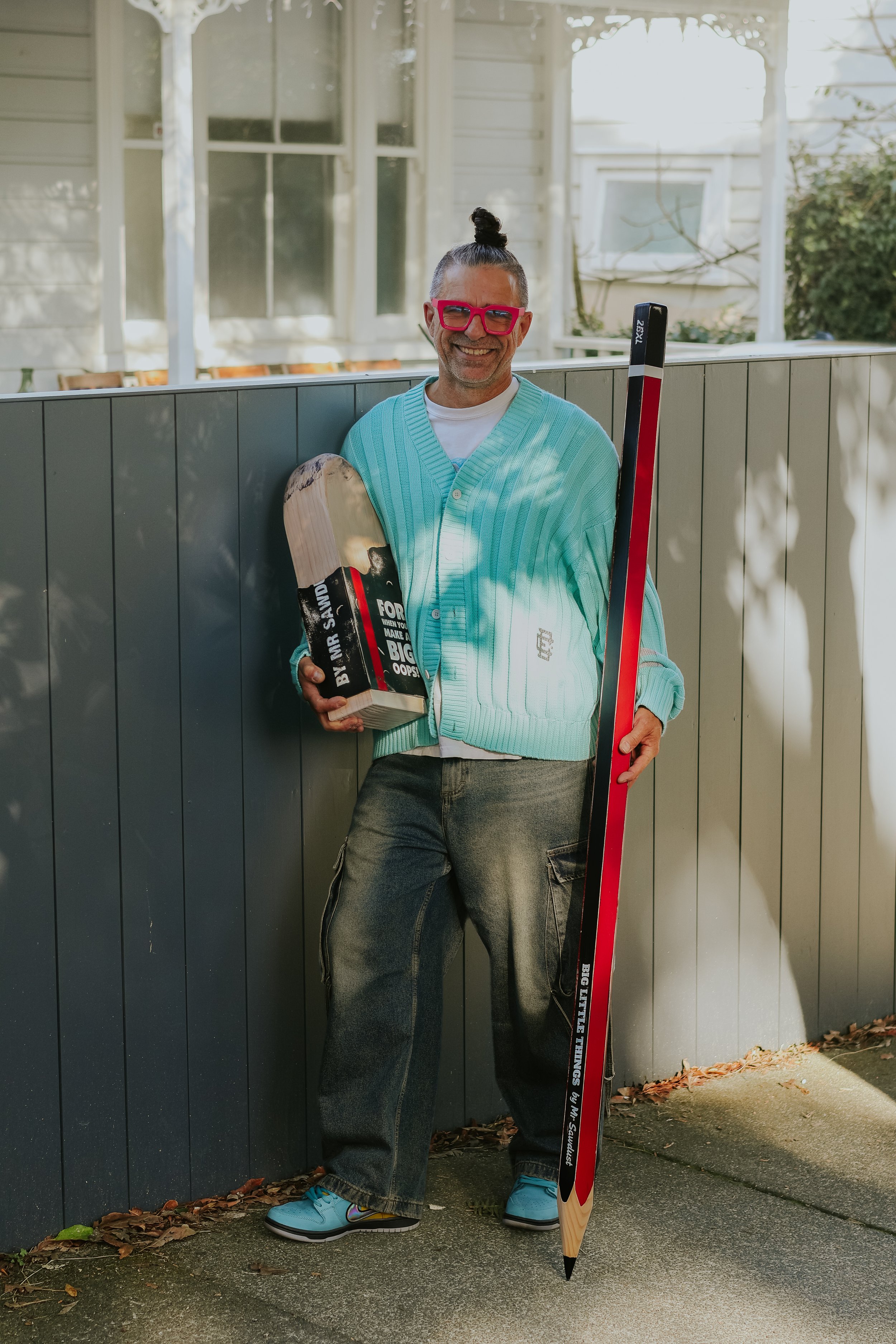 A man with a top knot hairstyle, pink glasses, and a turquoise cardigan smiling and holding a large wooden finger-shaped object and a hockey stick, standing outdoors near a house with siding and small windows.