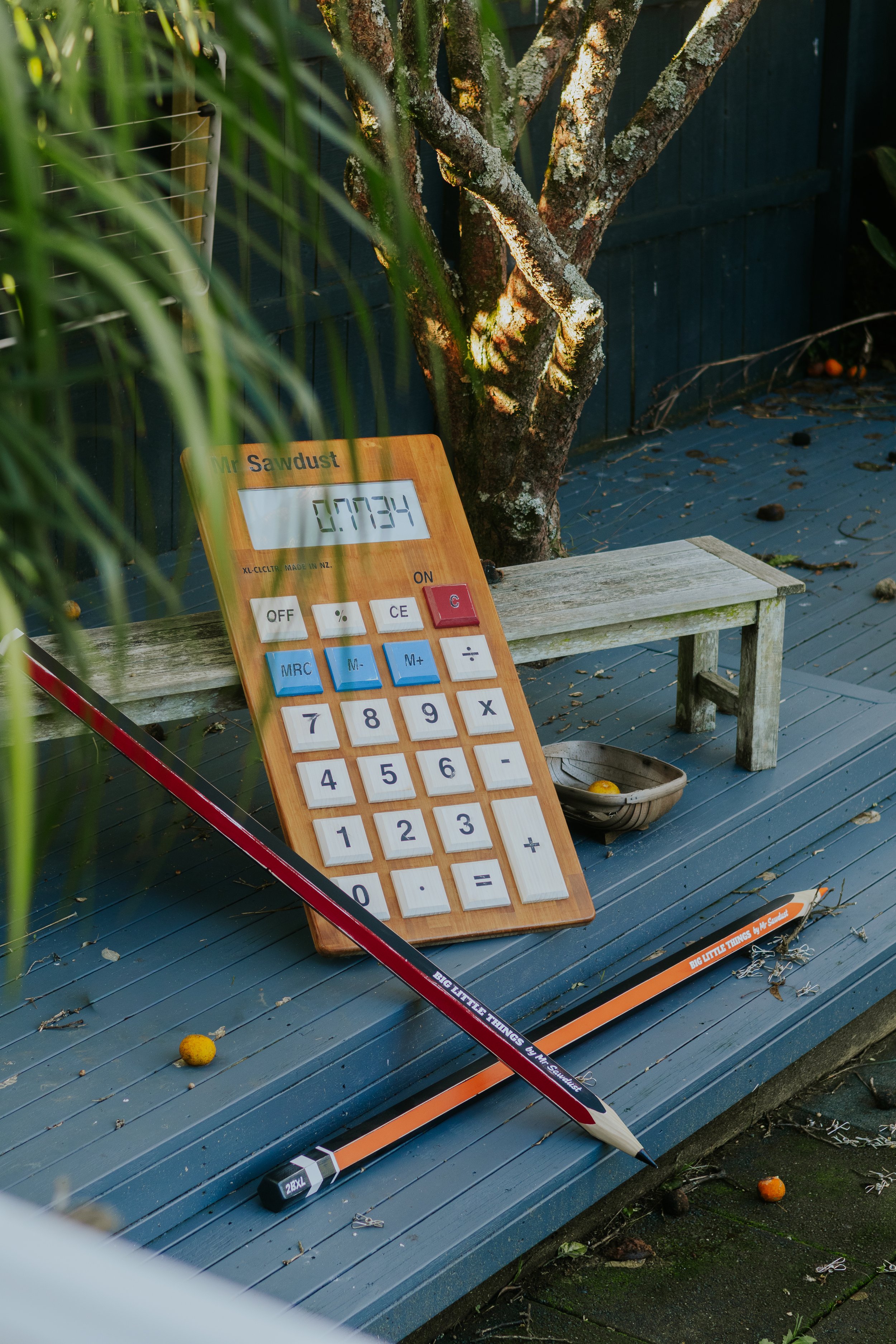 An outdoor scene with a wooden bench, a large calculator displaying 0.7734, a large model rocket, a broom, and a few scattered oranges and rocks on a blue wooden deck. A tree is also visible in the background.