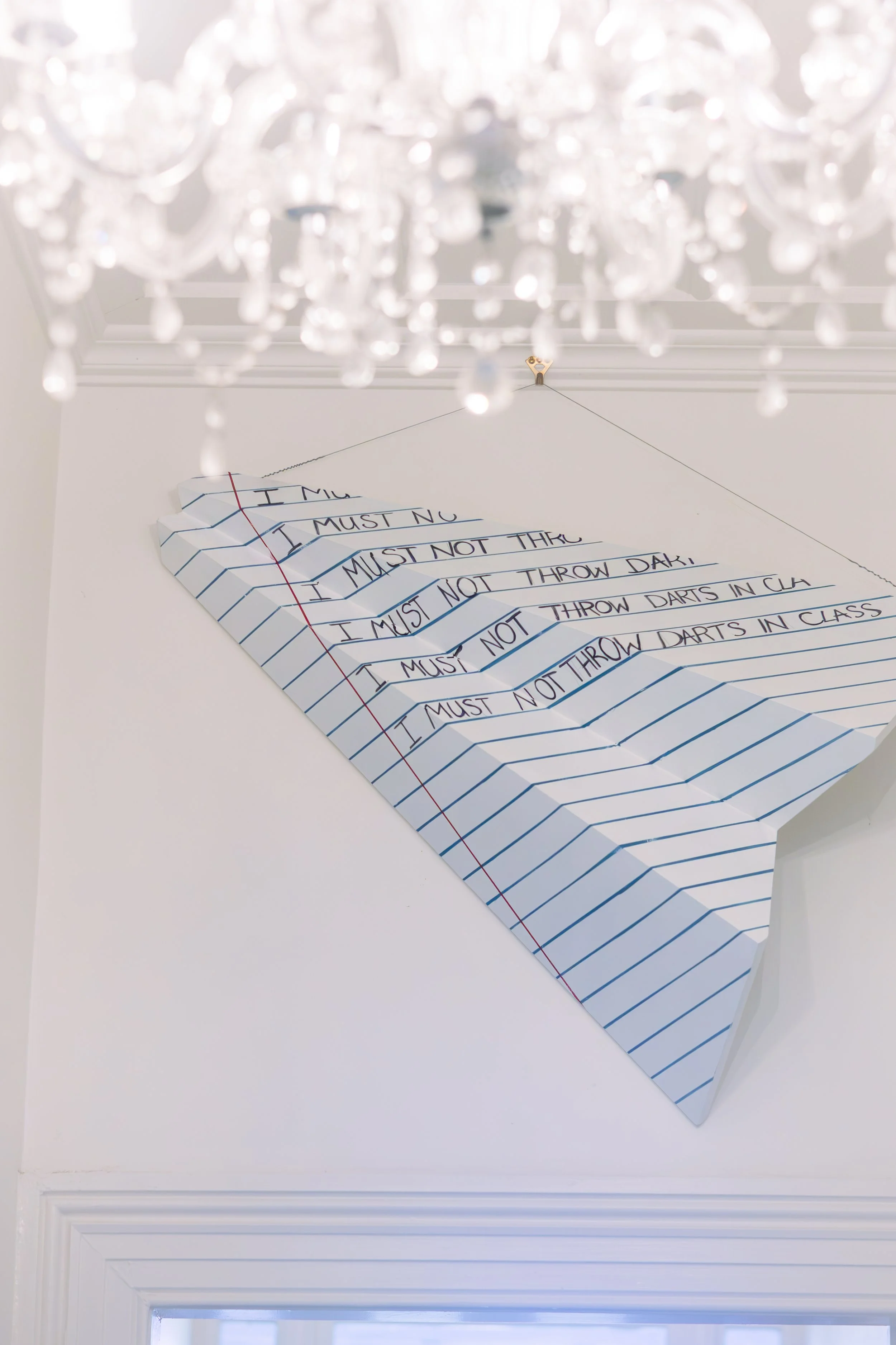 A chandelier hanging from the ceiling with a paper sign in the background that has handwritten text. The sign reads: 'I must not throw darts in class.'