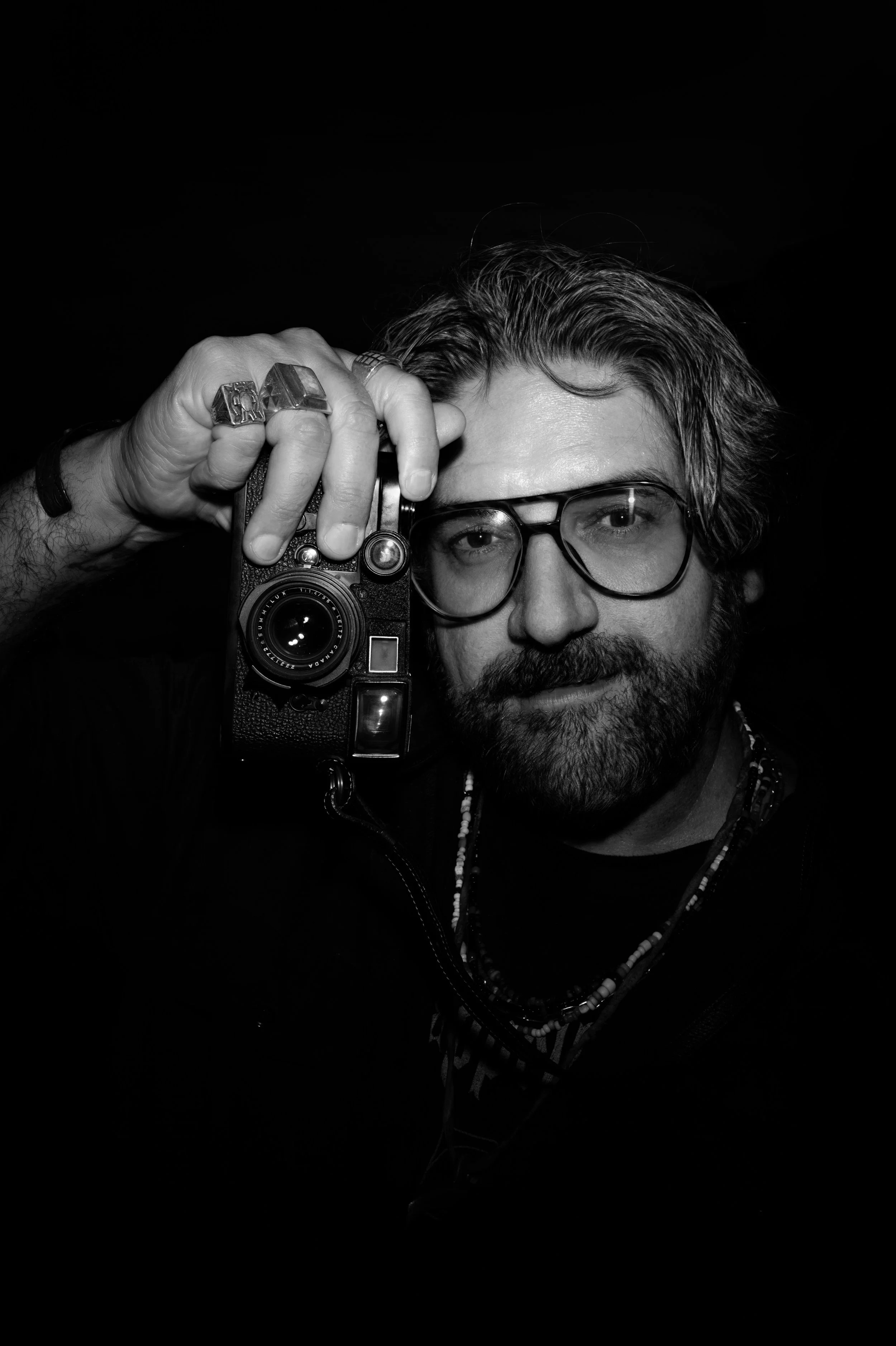 A man with glasses and a beard holding a vintage camera up to his face, wearing rings and necklaces, with a black background.