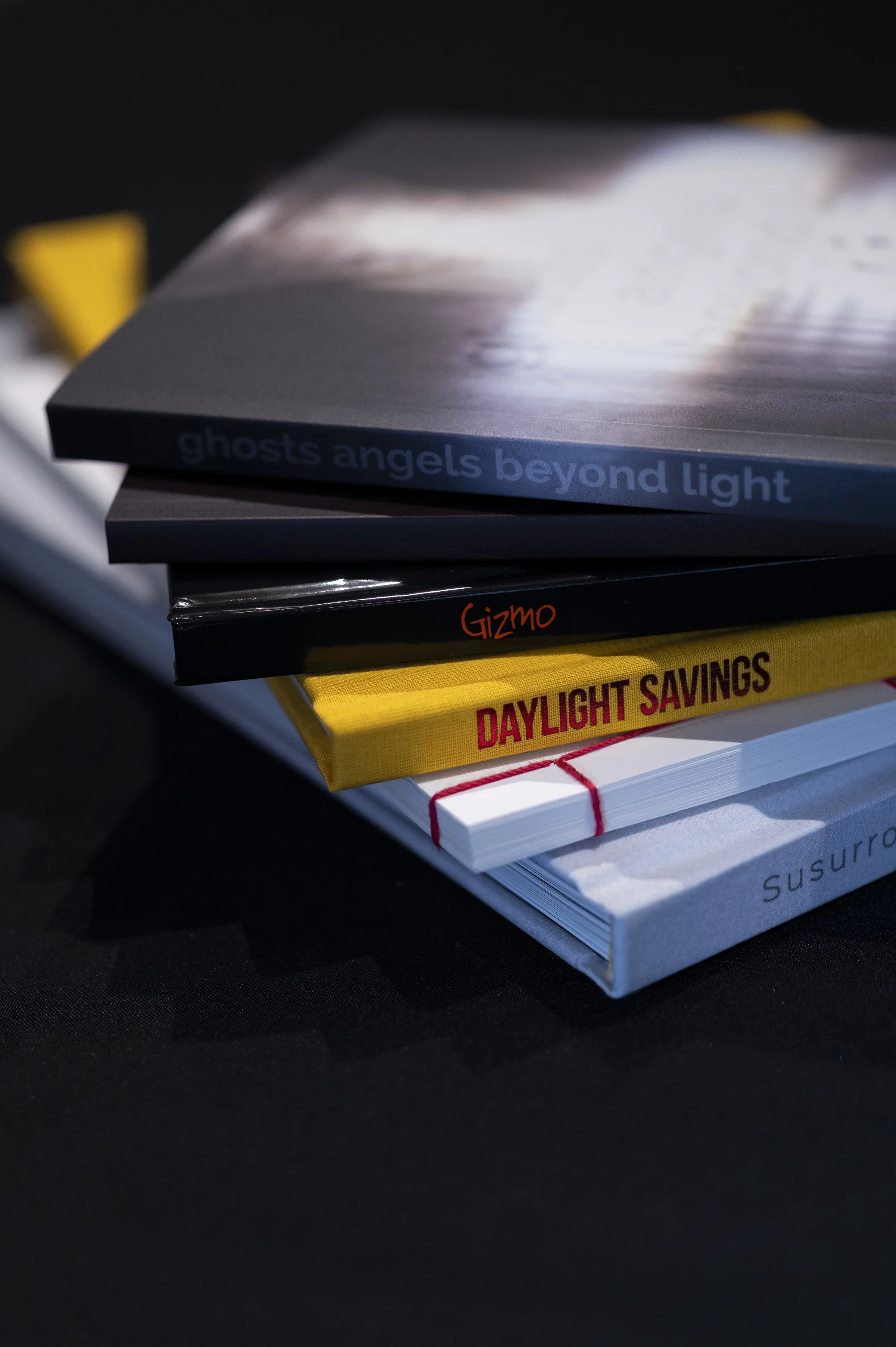 Close-up of a stack of books with titles visible, including 'ghosts angels beyond light', 'Gizmo', 'Daylight Savings', and 'Surgery'. The books are stacked on top of each other on a dark surface.