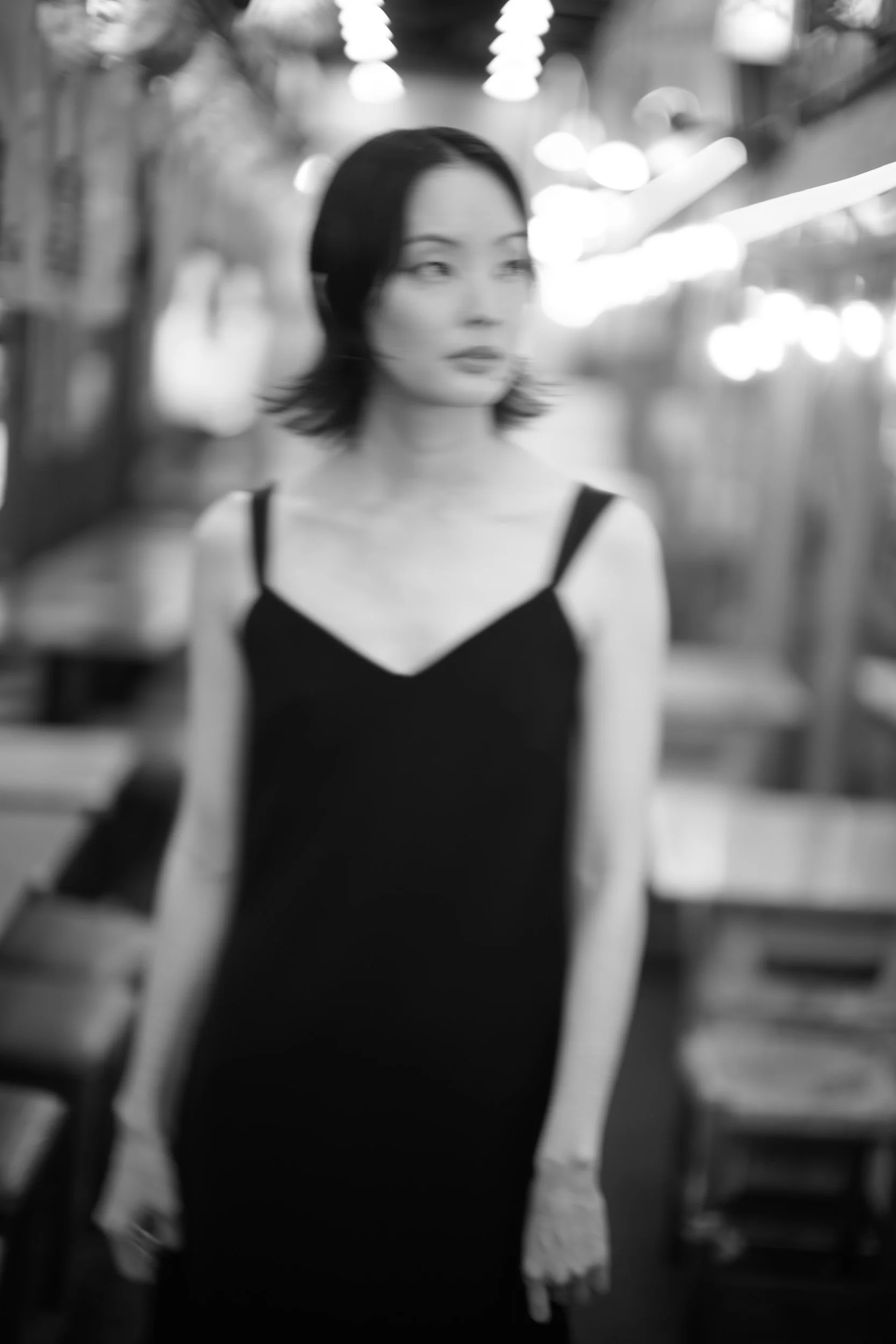 Black and white photo of a woman with short dark hair in an indoor setting, possibly a shopping mall or store, with blurred lights in the background.