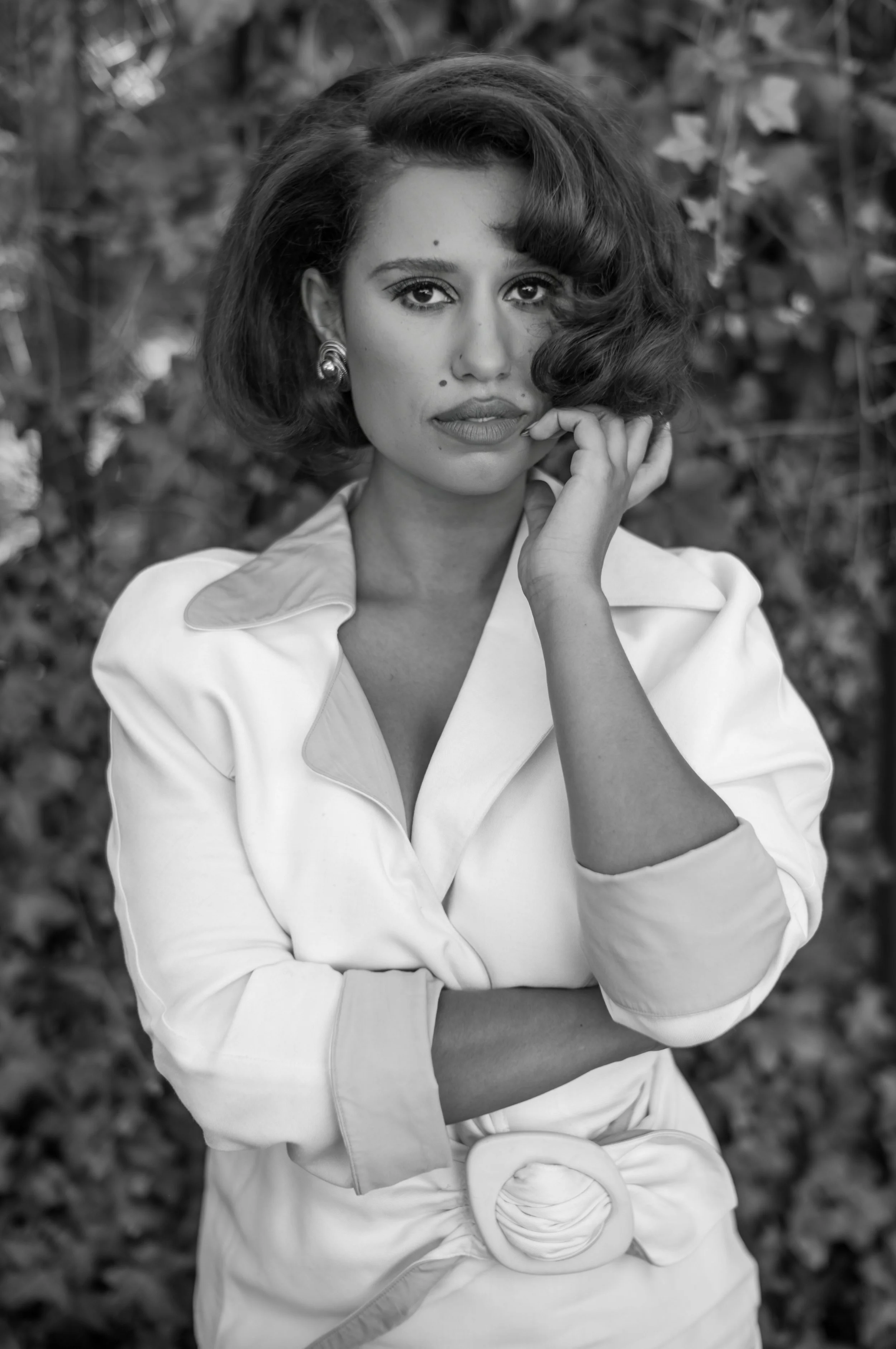 A woman with short, wavy hair, wearing earrings and a light-colored jacket, posing outdoors with a blurred leafy background. She has a calm expression and is looking directly at the camera.