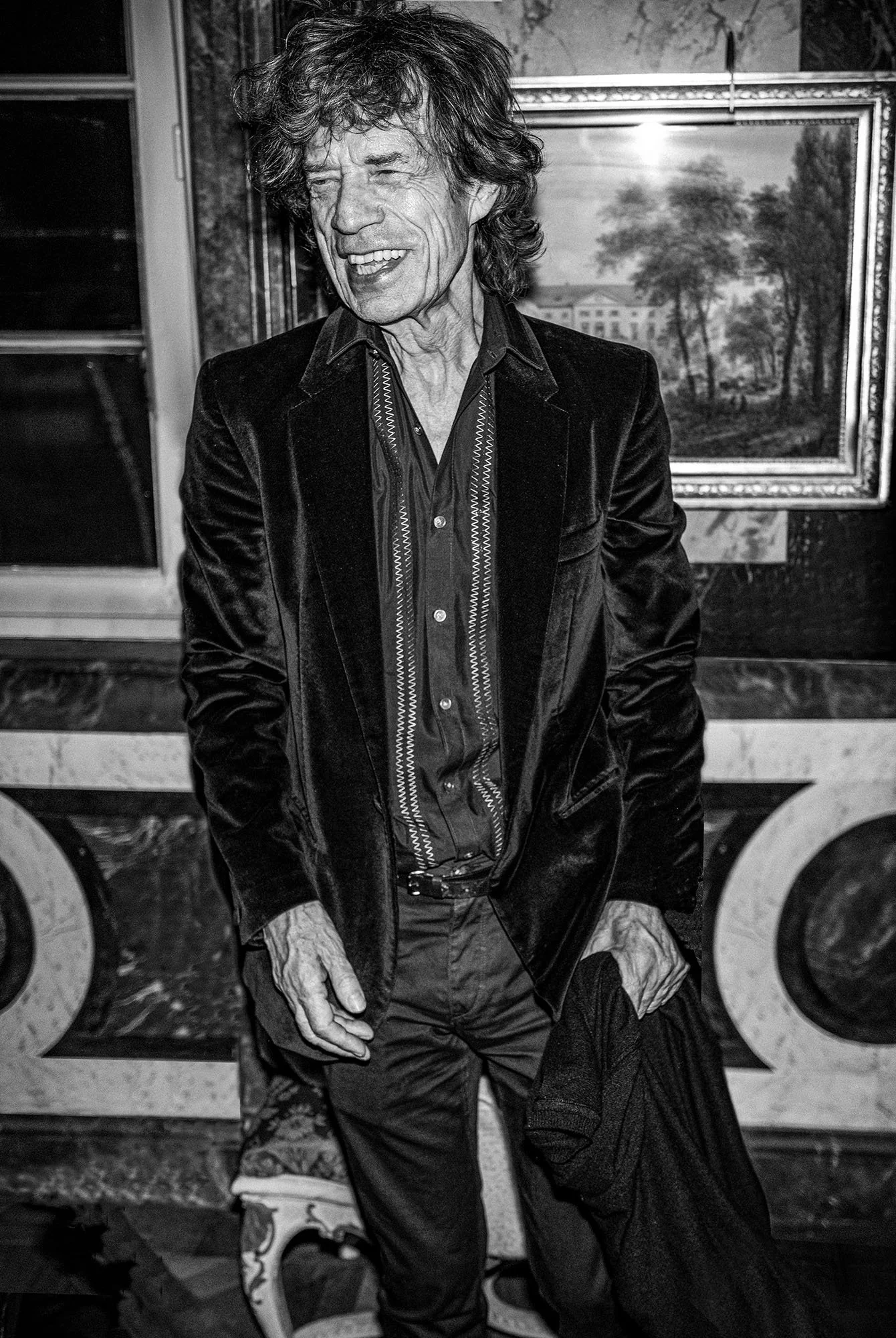 A black and white photo of a man with curly hair, smiling and wearing a velvet jacket over a button-up shirt, standing in front of a framed painting of trees and buildings.