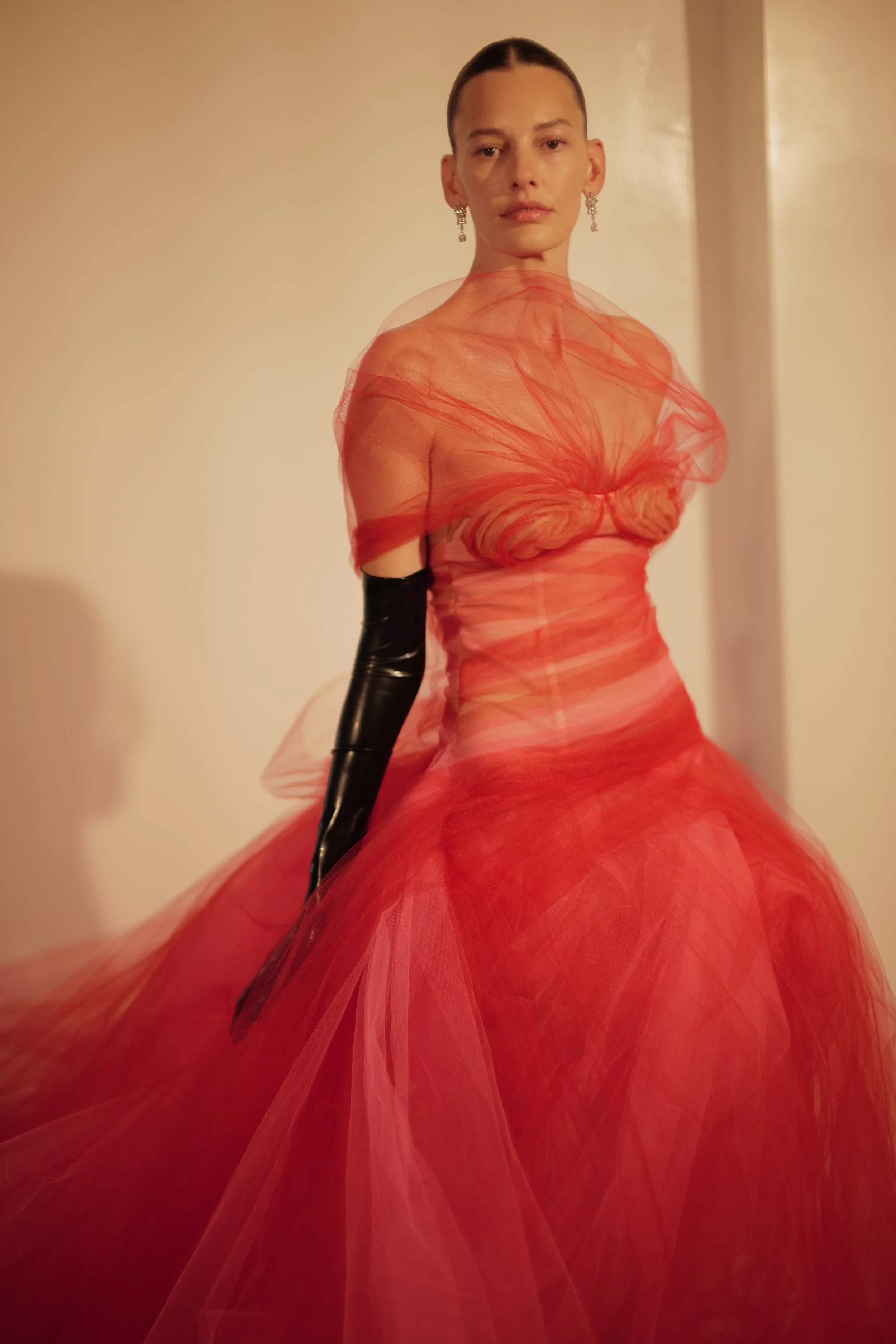 A woman with slicked-back hair in an elegant red gown made of tulle, wearing earrings and long black gloves, standing against a beige wall.