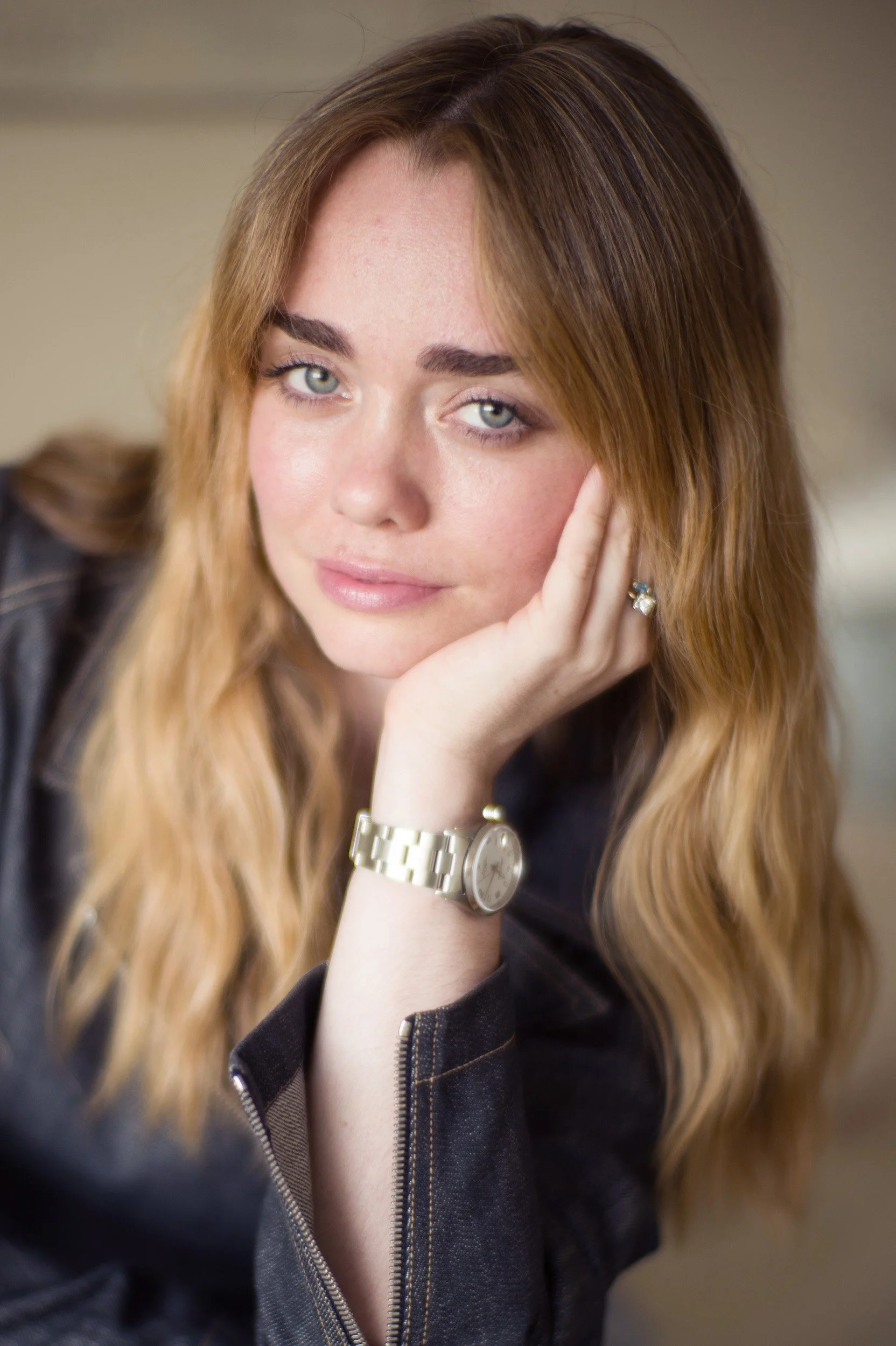 Close-up of a woman with long, wavy blonde hair, wearing a black jacket and a silver watch, resting her face on her hand with natural makeup and piercing blue eyes.