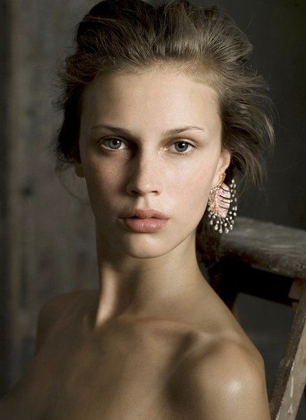 Close-up portrait of a young woman with fair skin, light brown hair, and wearing an earring, looking directly at the camera against a blurred background.
