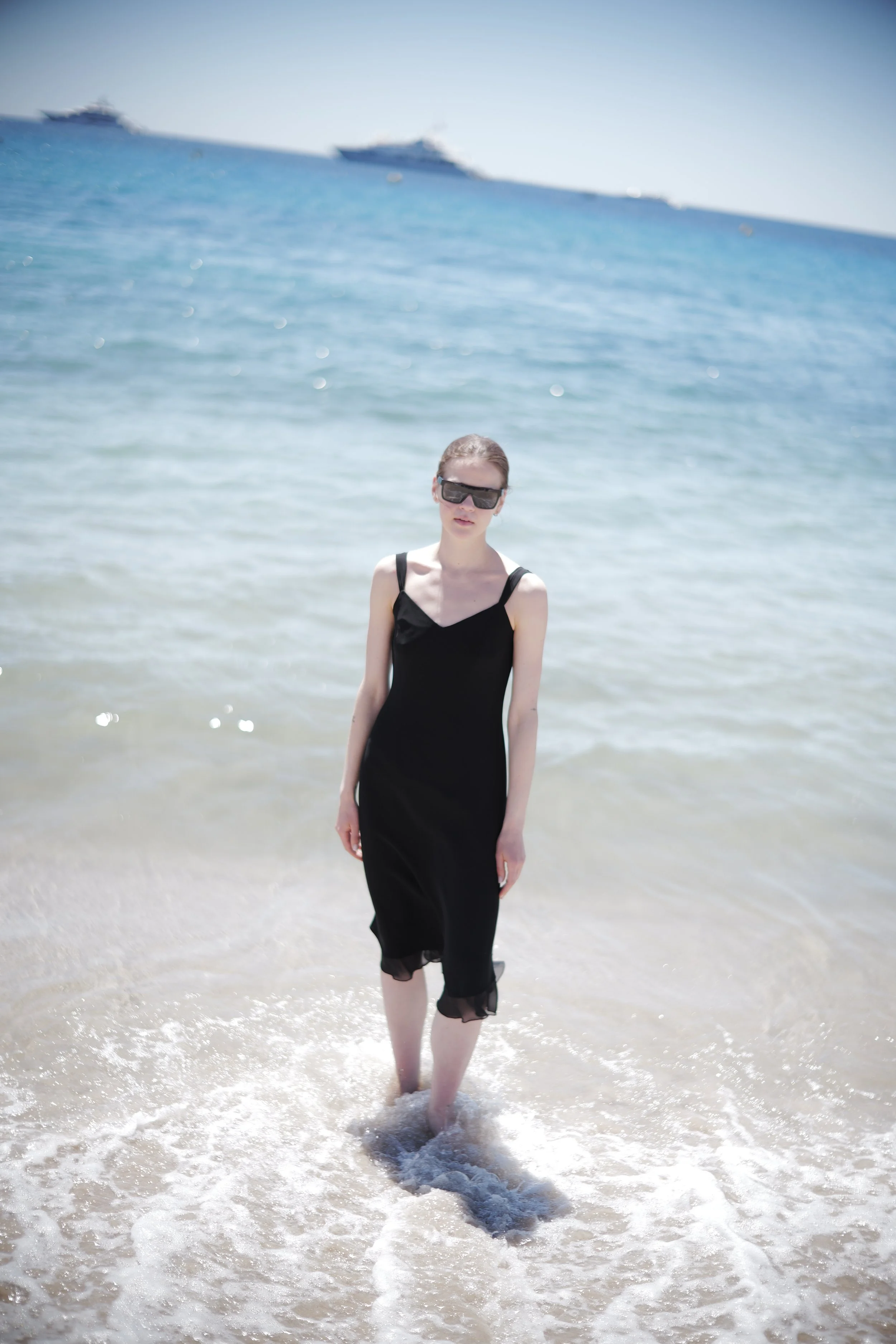 A woman in a black dress with sheer trim walking in the shallow surf at the beach, with boats visible on the horizon in the background.