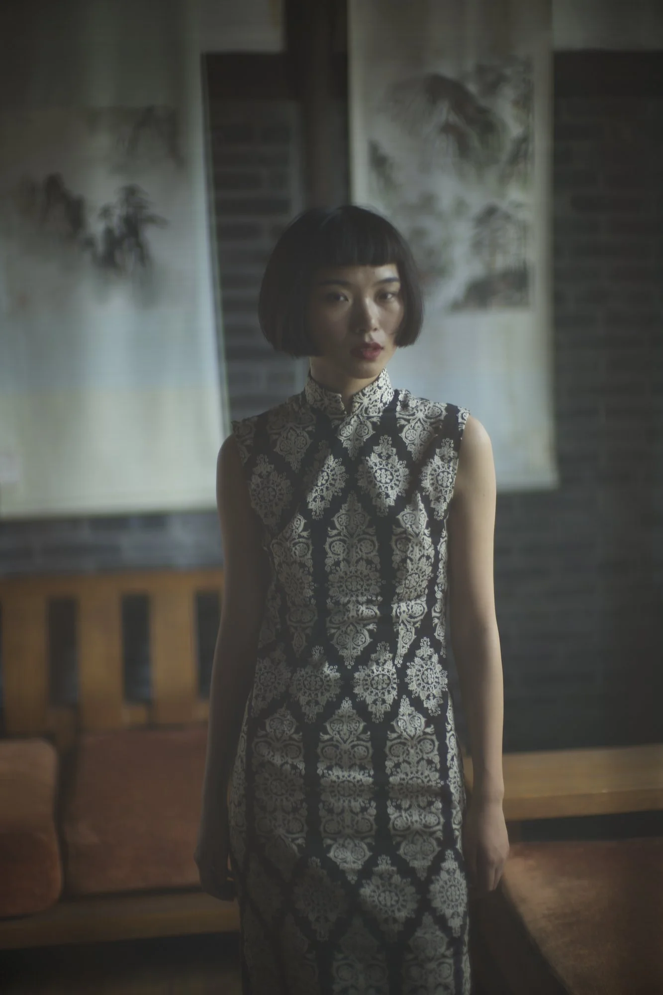 A woman with short black hair and bangs wearing a sleeveless black and white patterned dress standing in a room with traditional Asian paintings on the wall behind her.