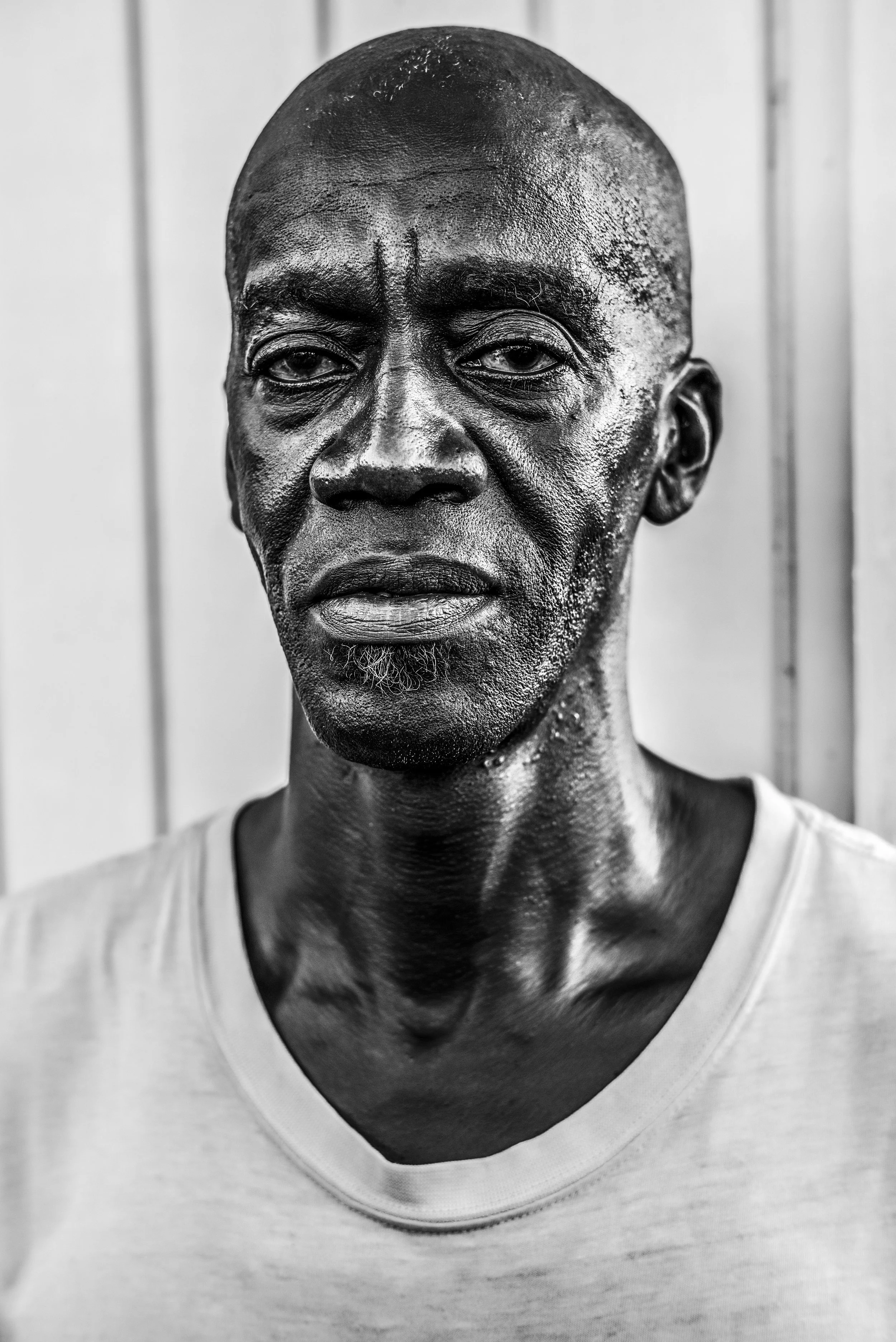 Close-up black and white portrait of a middle-aged African American man with a shaved head, wearing a white shirt, looking directly into the camera with a serious expression.