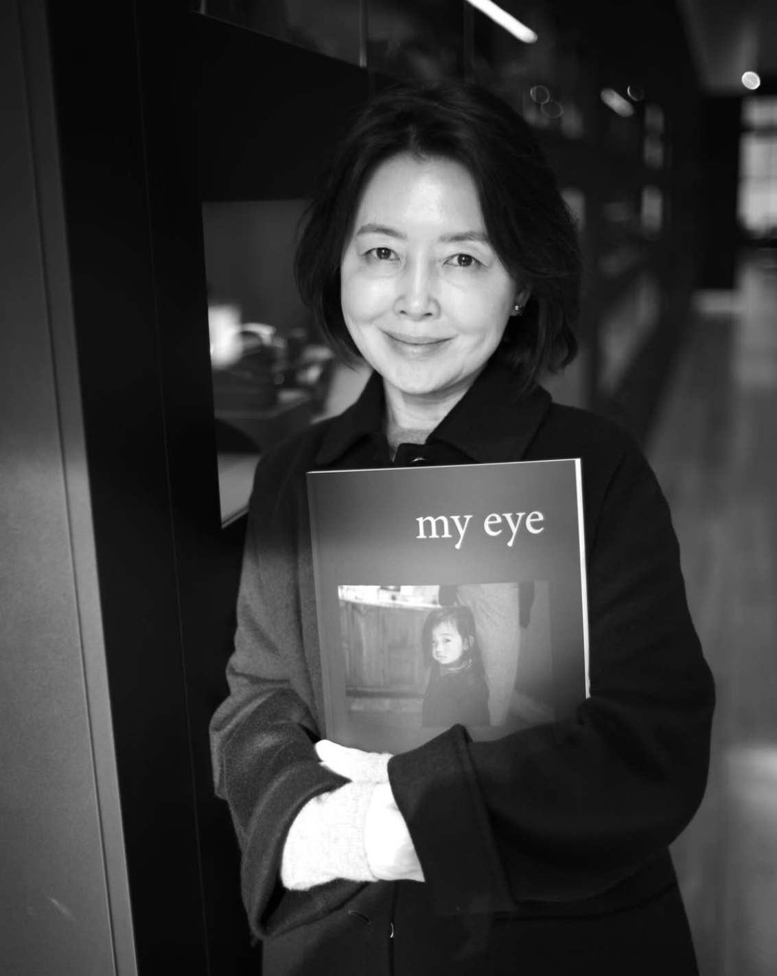A woman with short dark hair holding a magazine titled "my eye" featuring a child on the cover, smiling and looking at the camera.