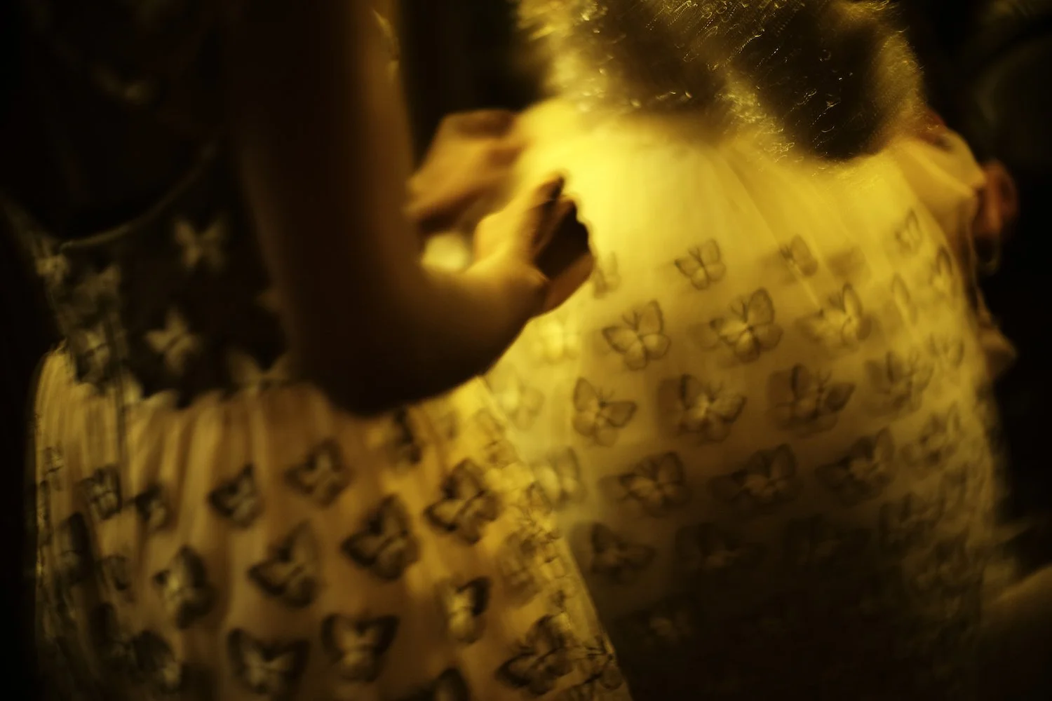 Close-up of a person with dark skin, wearing a yellow dress with butterfly patterns, illuminated by warm lighting.