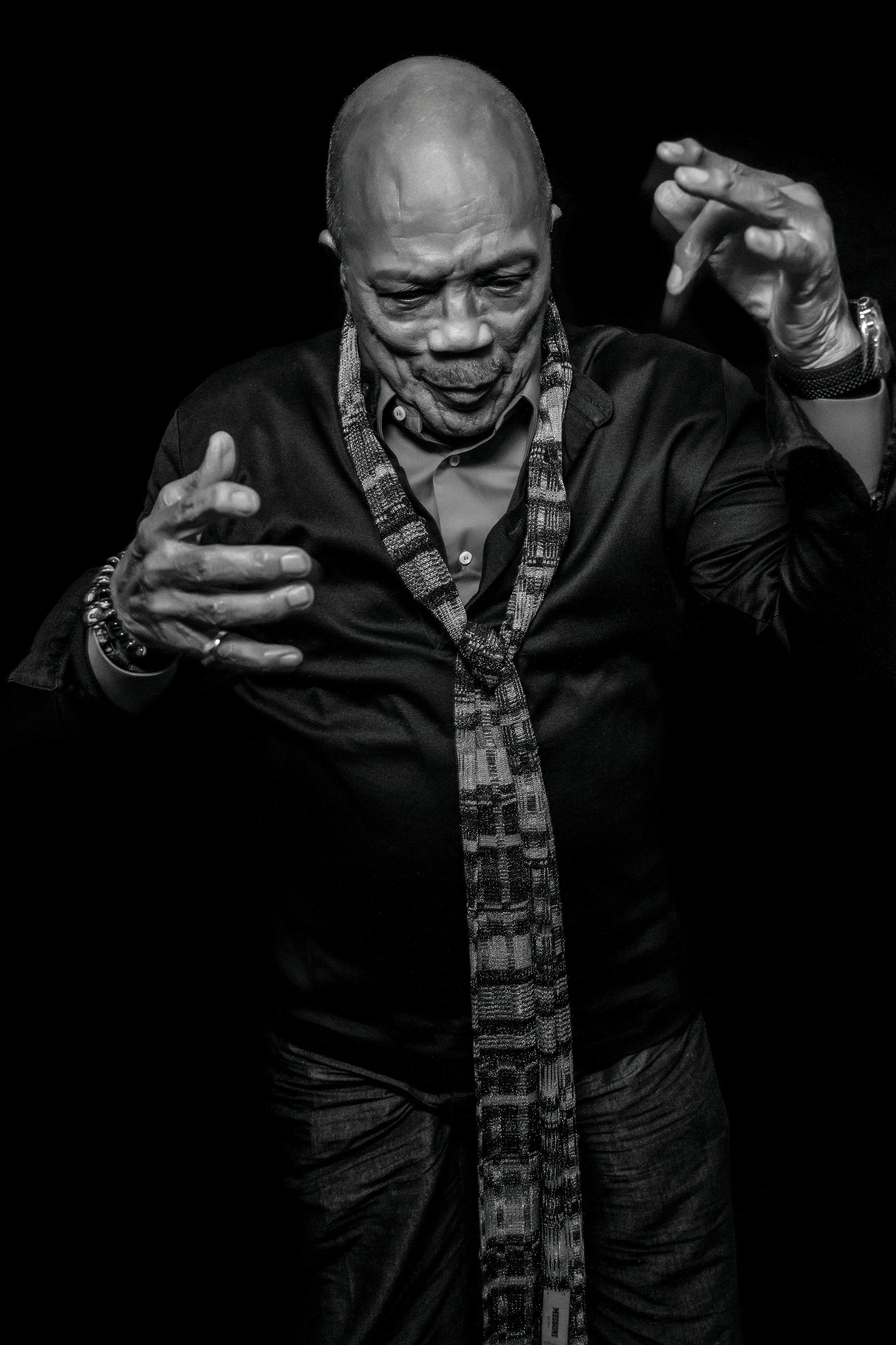 Black and white photo of a man with a bald head, wearing a dark shirt, a patterned scarf, and various bracelets, making expressive hand gestures against a dark background.