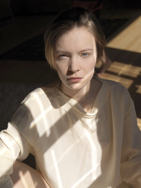A young woman with short brown hair and blue eyes looking directly at the camera, wearing a cream-colored shirt with sunlight casting patterns on her face and shirt.