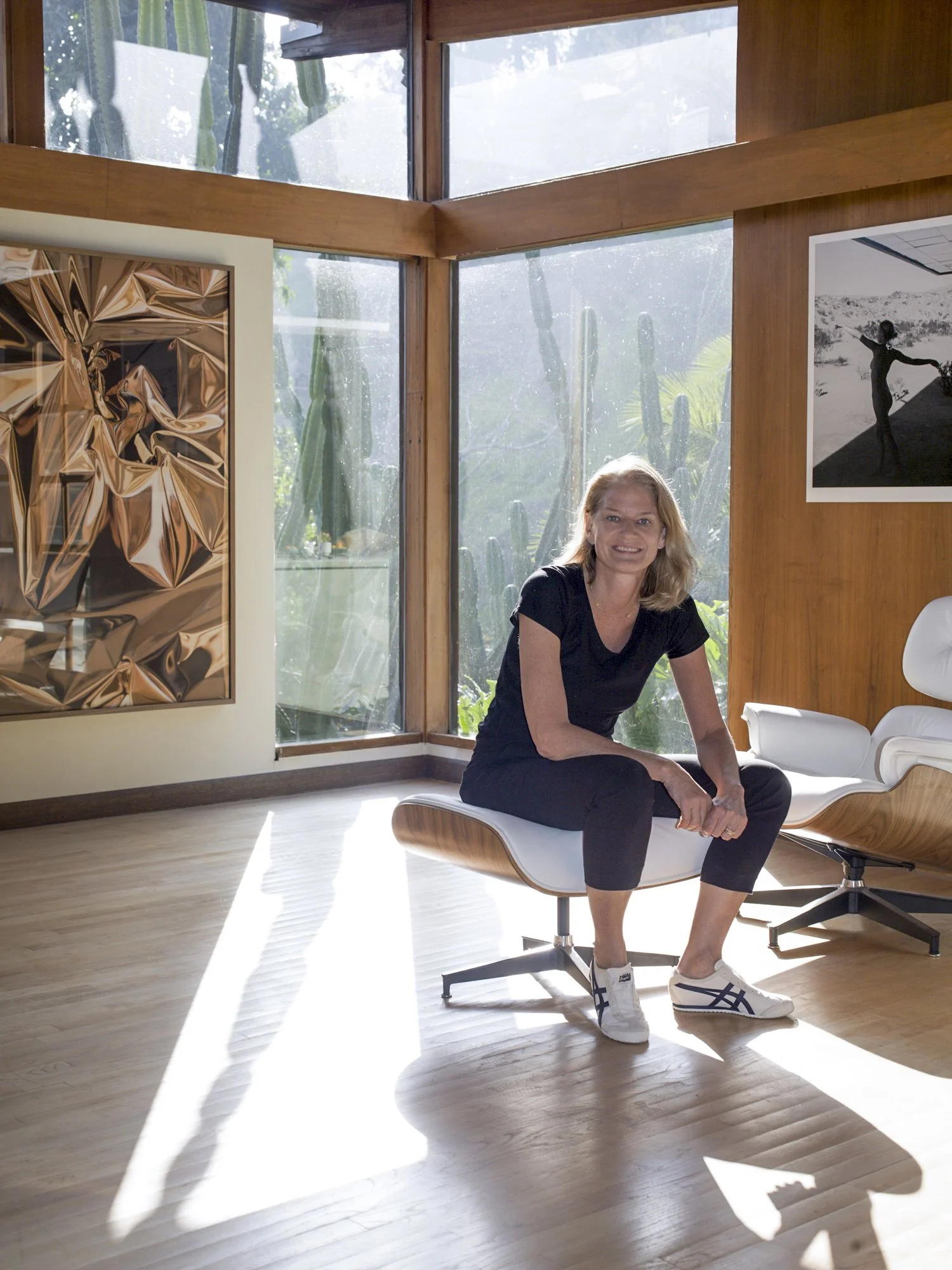 Woman with blonde hair sitting on a modern wooden and white chair inside a room with large glass windows showing cactus plants outdoors.