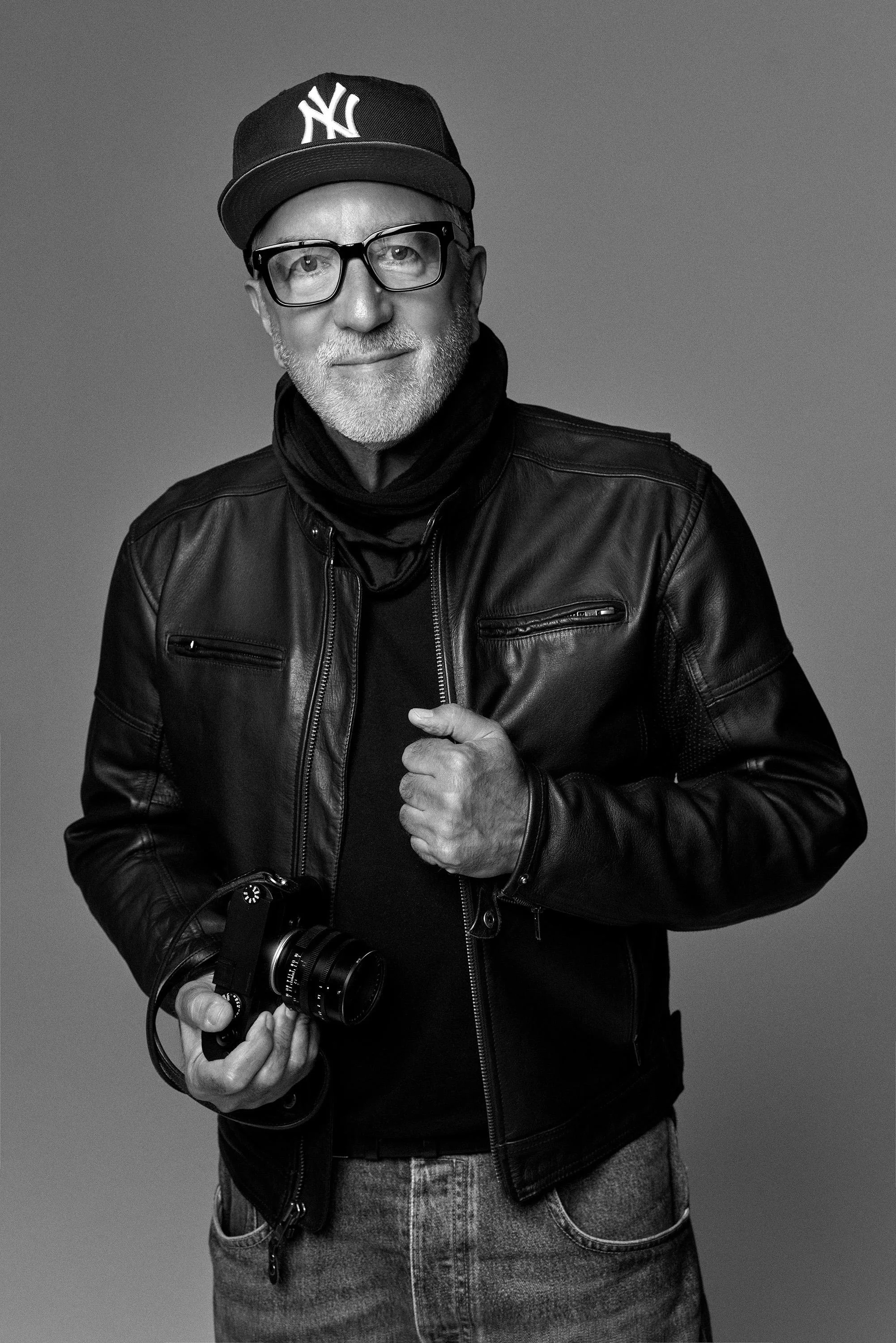 Black and white portrait of an older man wearing a New York Yankees cap and glasses, holding a camera in his right hand, dressed in a leather jacket and jeans, with a neutral background.
