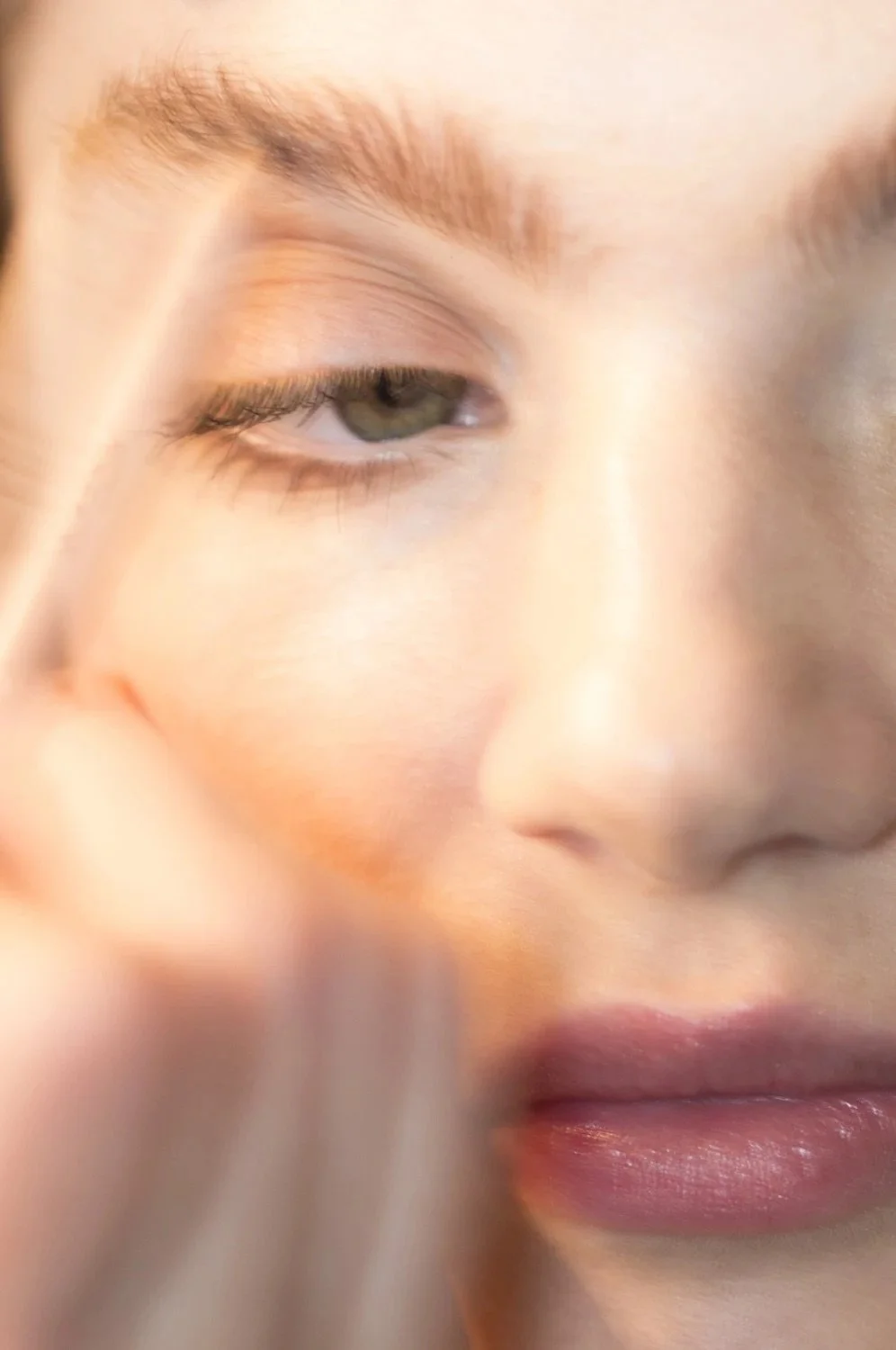 Close-up of a person's face with one eye partially closed, light skin, brown eyebrows, and light makeup, with a soft focus on the lips and skin.