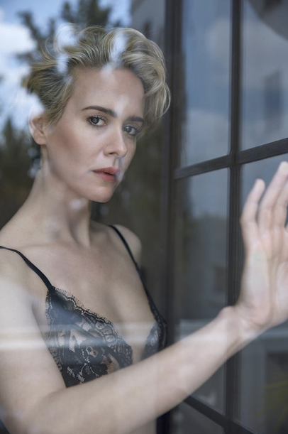 A woman with short, wavy blonde hair and a serious expression, wearing a black lace top, standing by a window with her hand on the glass.