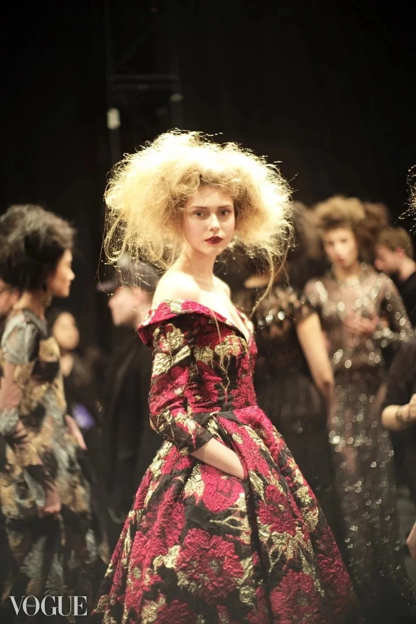 A woman with curly blonde hair wearing a floral red and black gown with pockets, standing on a fashion runway. Other models in ornate dresses are blurred in the background.