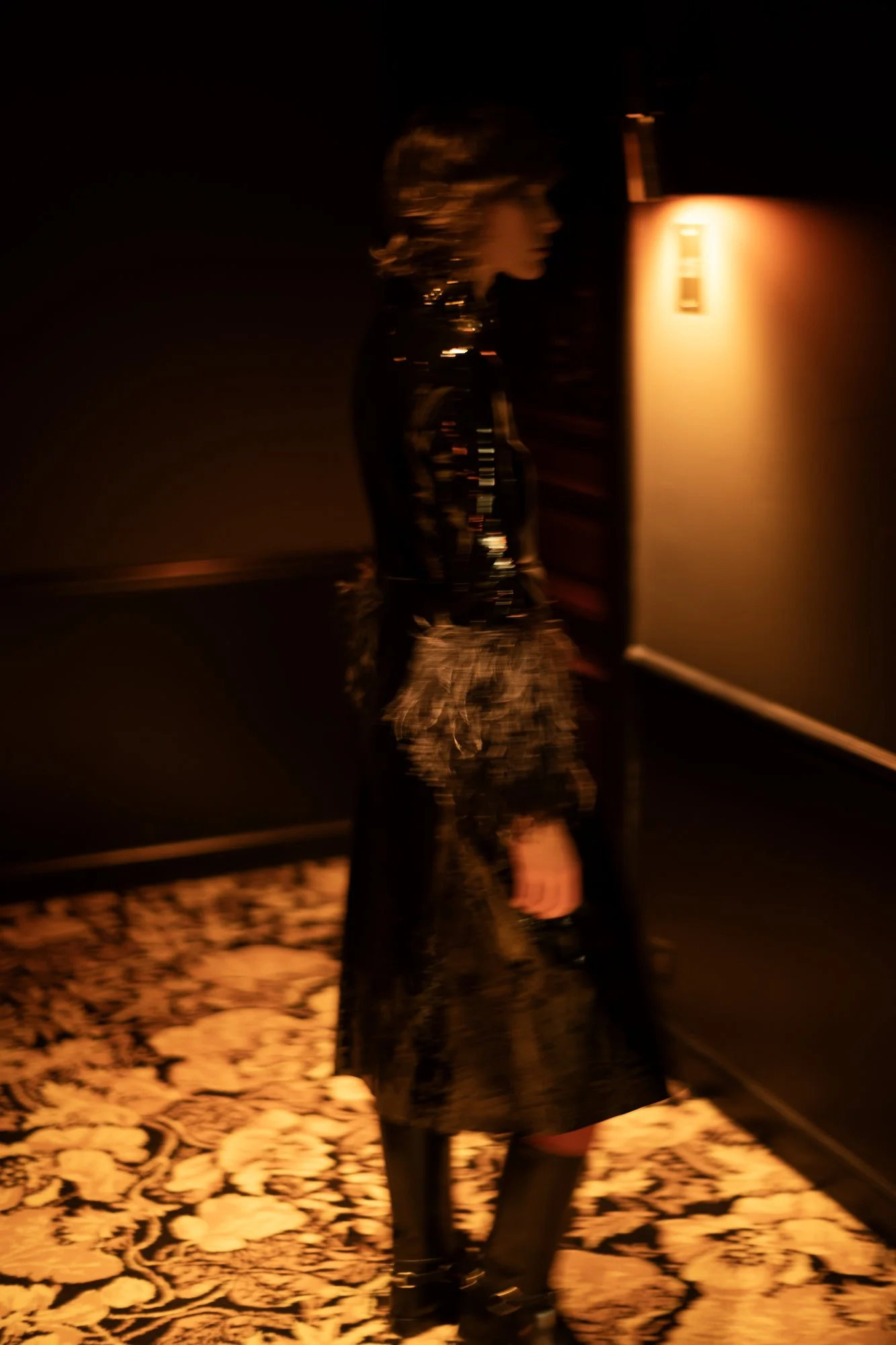 A woman standing in a dimly lit room near a wall with a warm light, wearing a dark patterned outfit and black boots, with a patterned carpet on the floor.