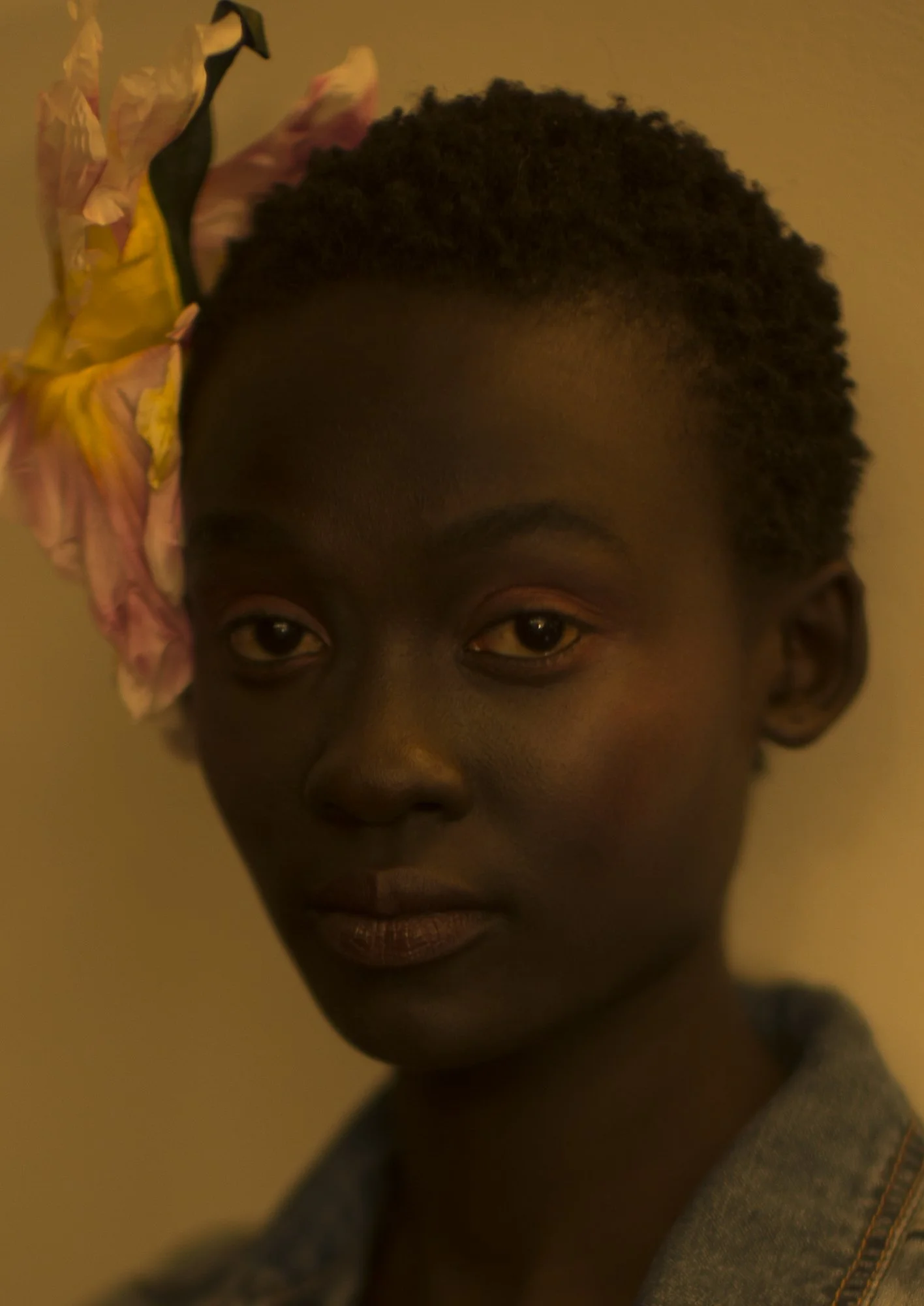 Close-up of a young person with dark skin and short curly hair, wearing a piece of floral fabric on their head and a denim jacket.