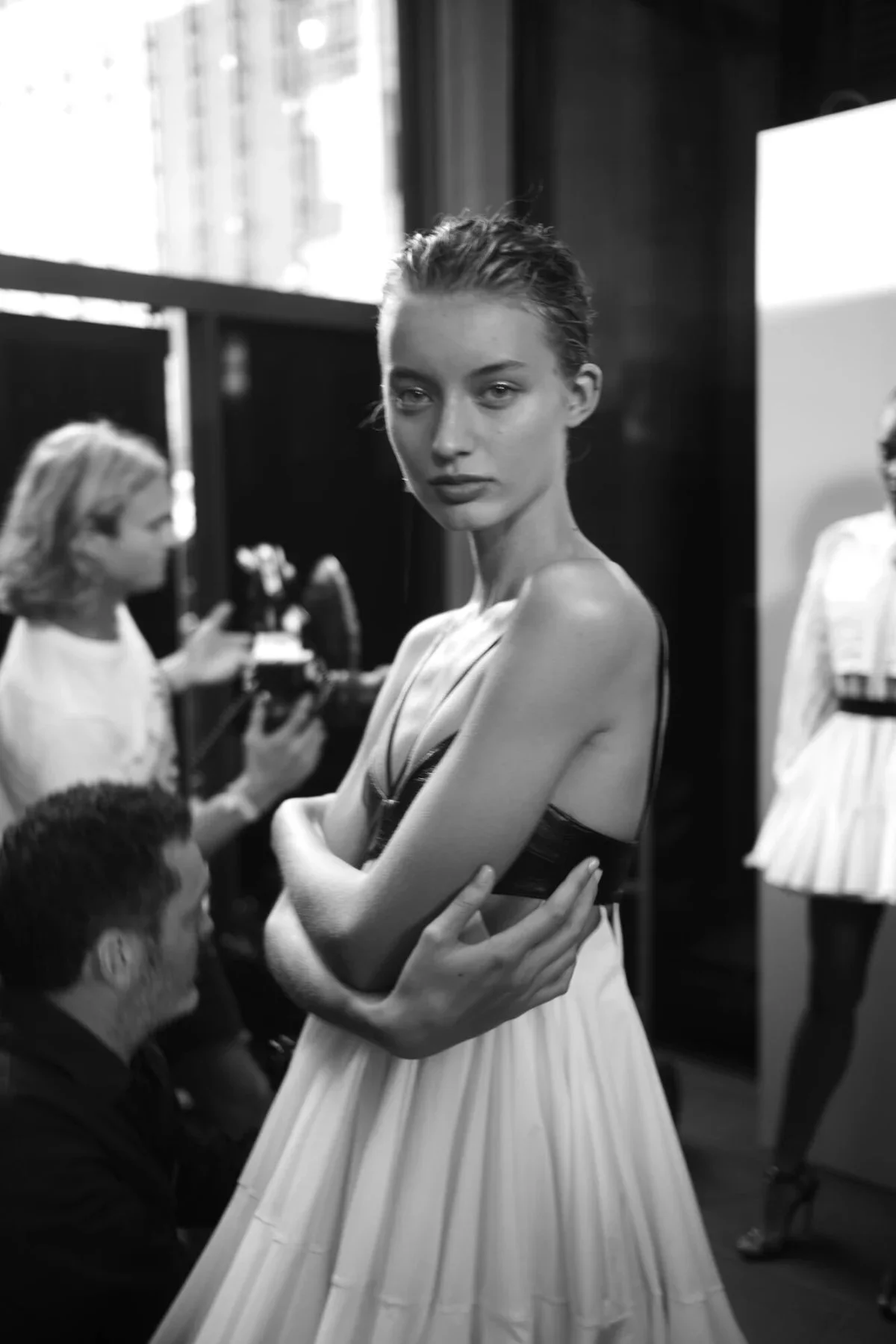 Black and white photo of a model with crossed arms and a serious expression, surrounded by people and clothing mannequins in a backstage or studio setting.