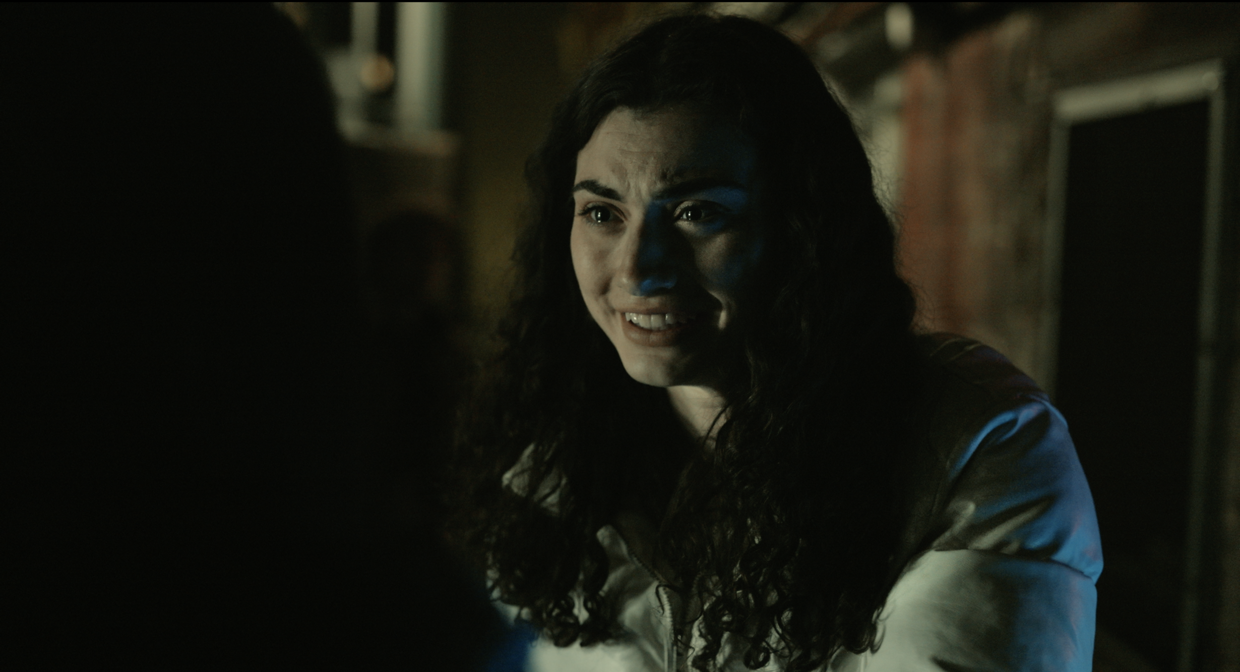 A woman with long dark curly hair smiling in a dimly lit room.