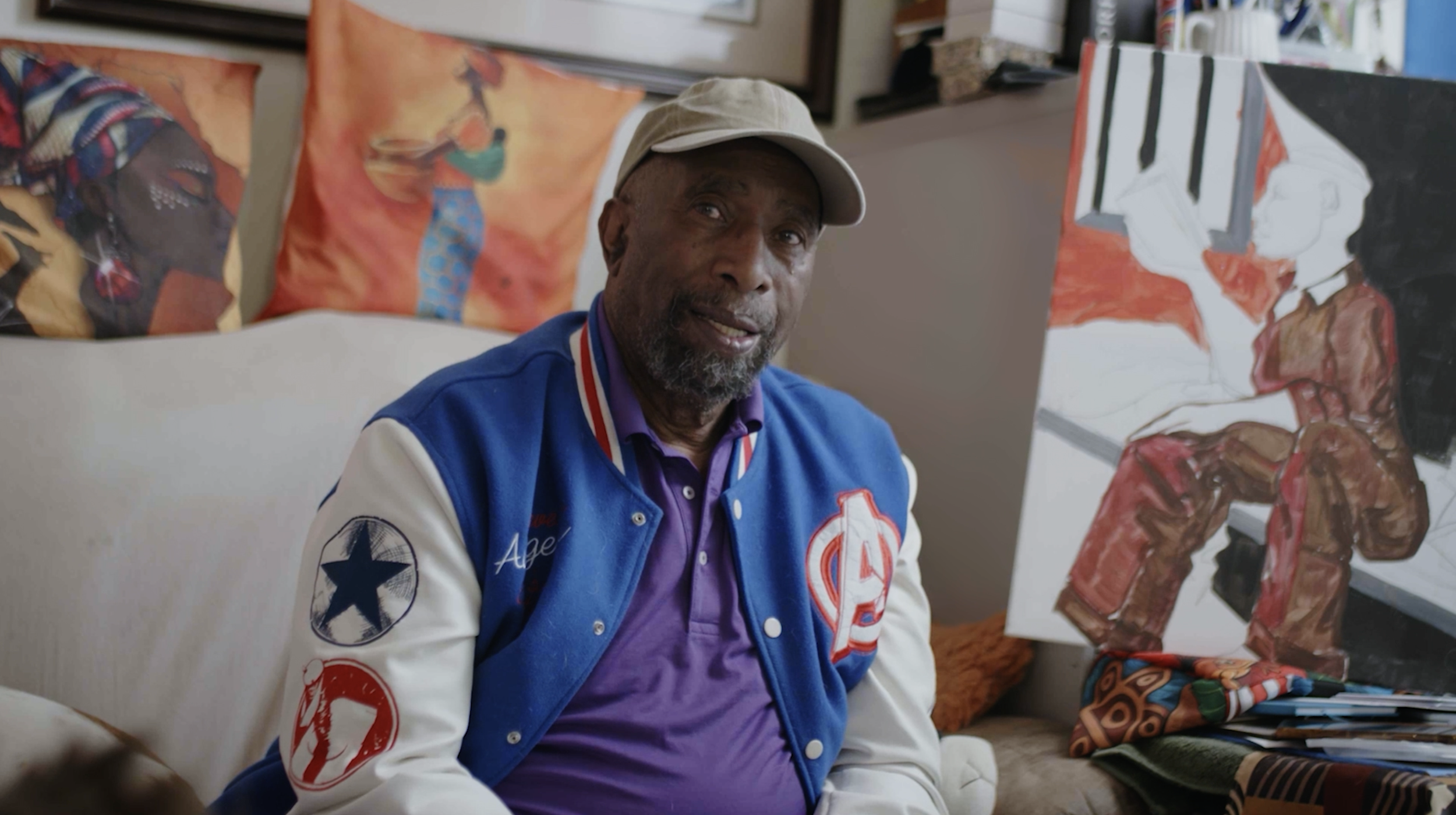 An older man with a beard wearing a beige cap and a colorful varsity jacket with patches, seated in front of a colorful abstract painting in an art studio.