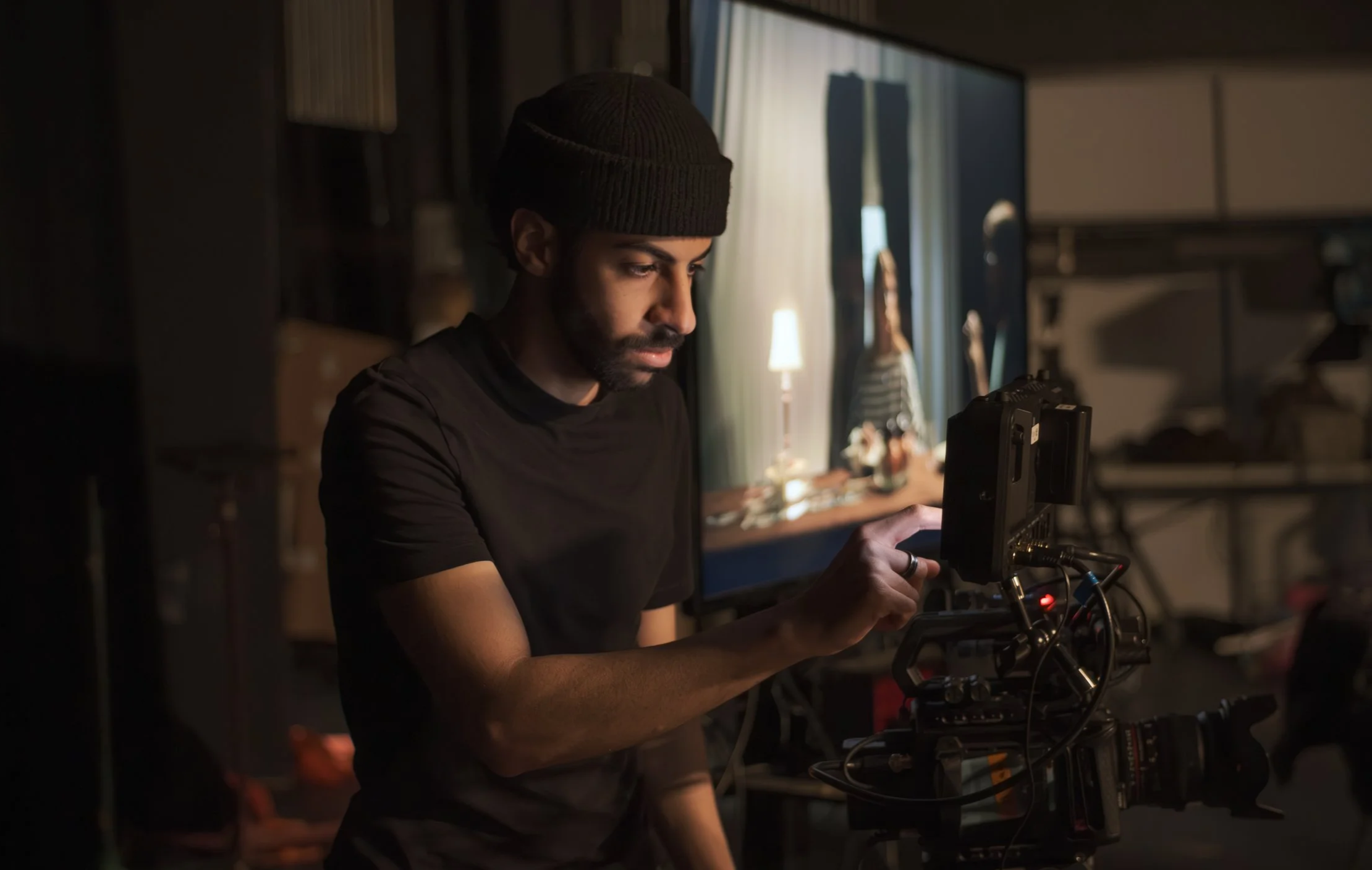 A person wearing a dark beanie and black T-shirt working with electronic equipment in a dimly lit room.