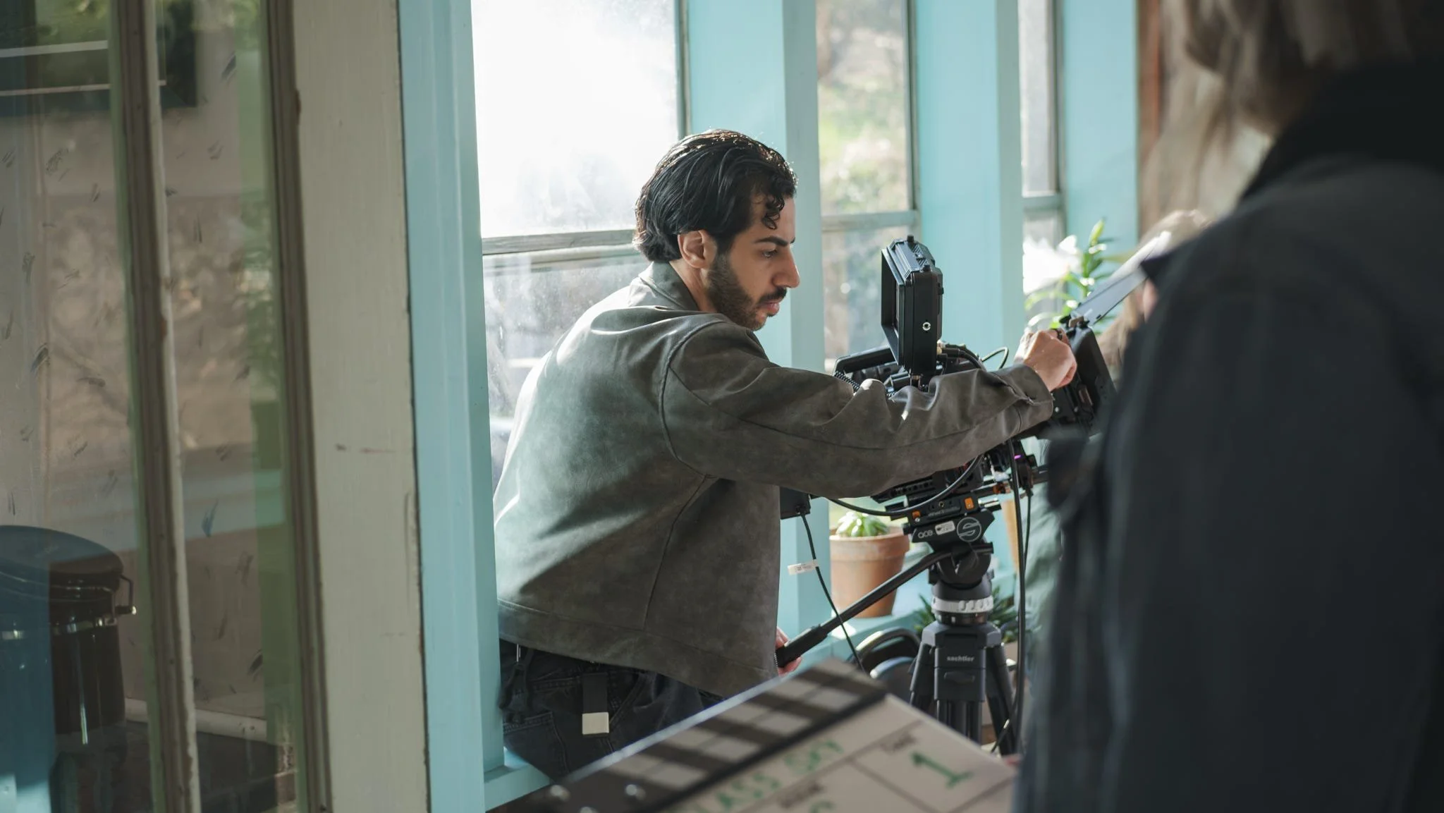 A man operating a professional camera mounted on a tripod, adjusting settings, while another person stands nearby inside a room with large windows and green plants.