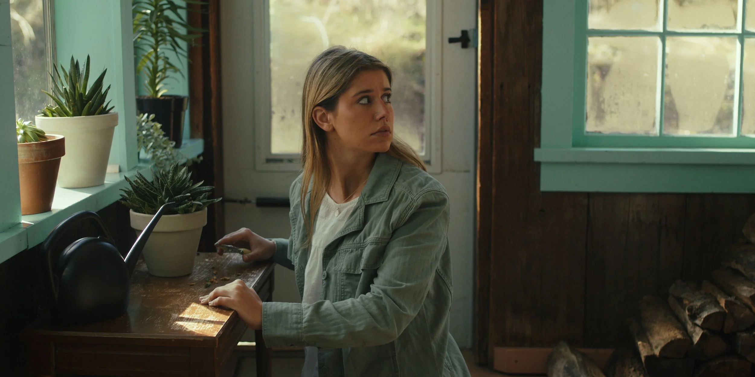 A woman with blonde hair, wearing a green jacket and white shirt, sitting at a wooden table in a room with potted plants on the windowsill, looking to her right with a serious or concerned expression.