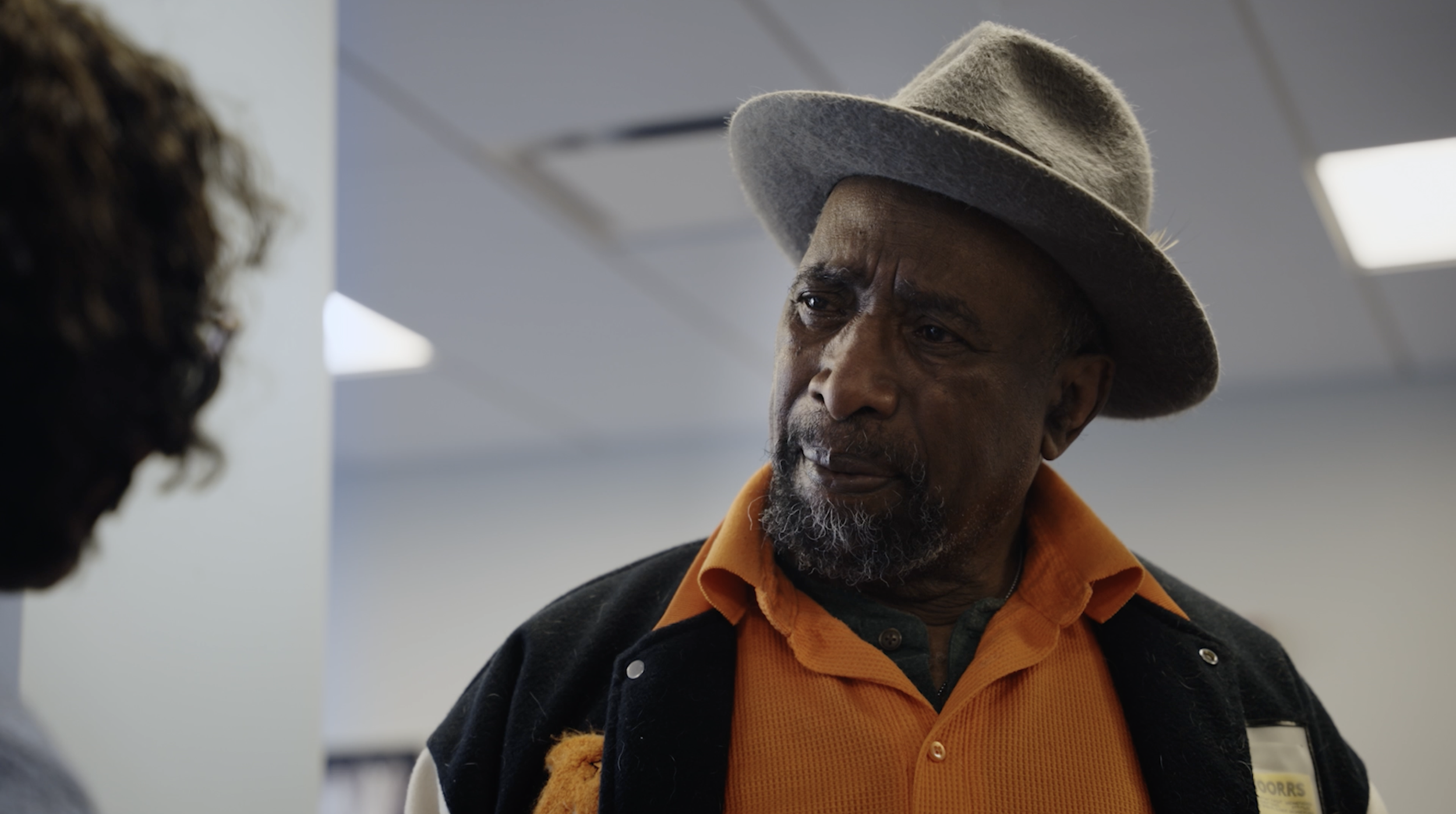 An older man wearing a light gray fedora hat, orange shirt, and black jacket engaged in conversation with a woman whose curly hair is visible on the left side of the image. The setting appears to be indoors, possibly in an office or public building.