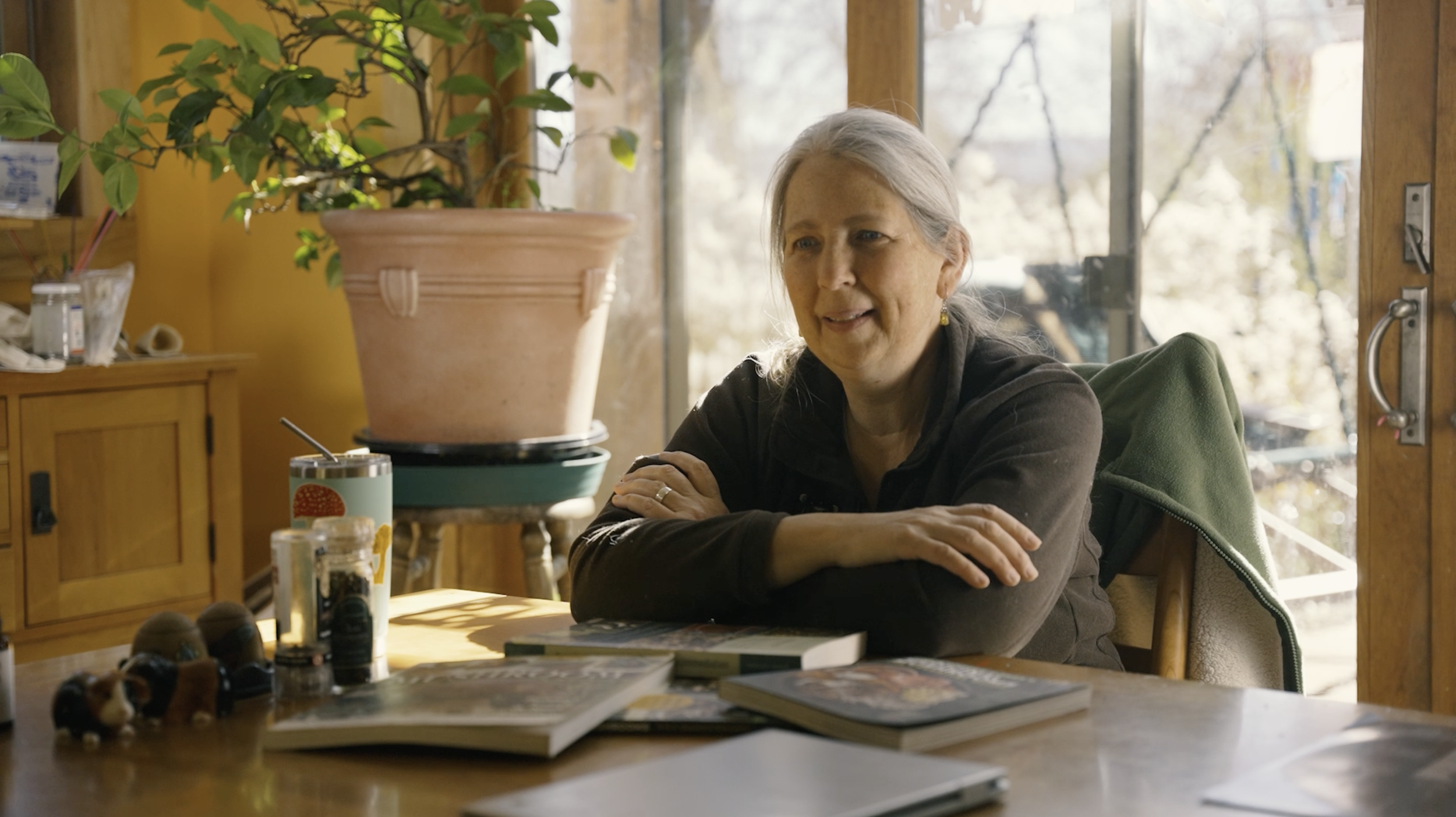 An elderly woman with gray hair sitting at a wooden table in a kitchen or dining area, smiling and resting her arms on the table. There is a large potted plant on the table, various jars, books, and small objects on the table and a cabinet in the background. Natural light is coming through large windows.