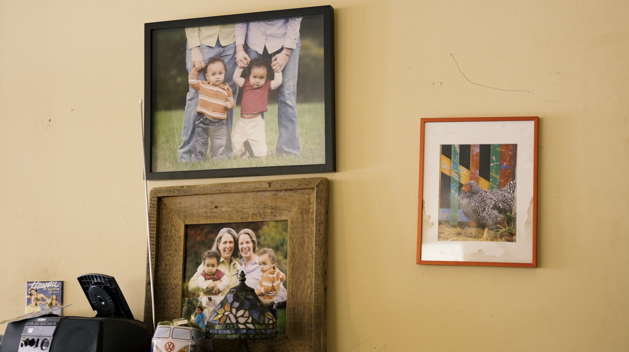 A wall with three framed pictures, one of two children with two adults, another of two women with children, and a photo of a bird and colorful background. A radio, a small decorative lamp, and a Volkswagen model are also visible on a table.