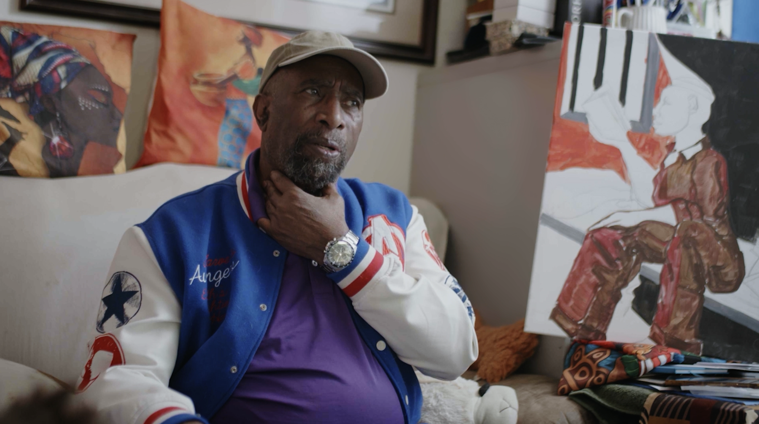 An older man with a beard wearing a beige cap, colorful varsity jacket, and purple shirt, sitting on a couch in an art studio. He is touching his neck and looking thoughtfully at a large painting of a woman with long dark hair and a red outfit.