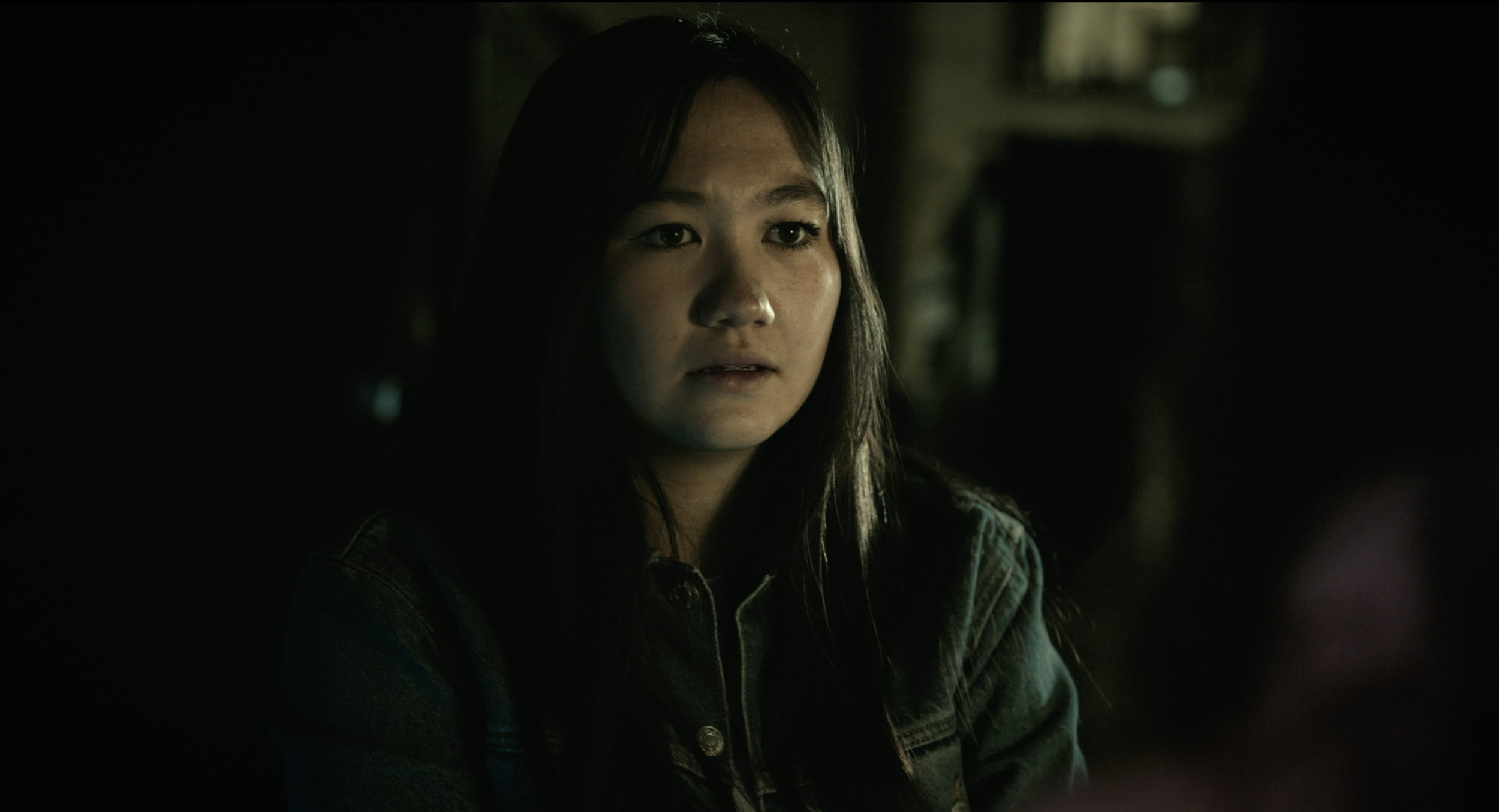 A young woman with long dark hair and light makeup looking slightly to the side, in a dark indoor setting.