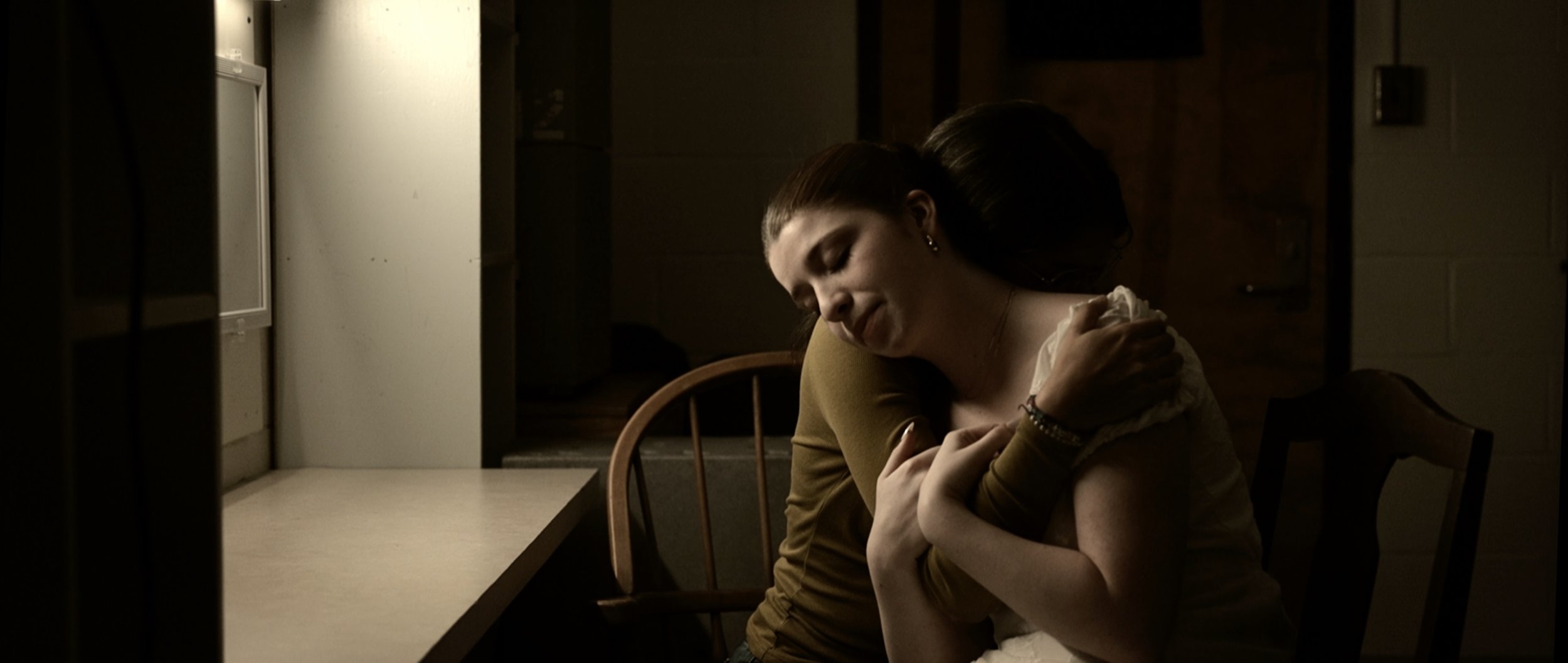 Two women hugging each other in a dimly lit room, one with brown hair and the other with dark hair, embracing warmly.