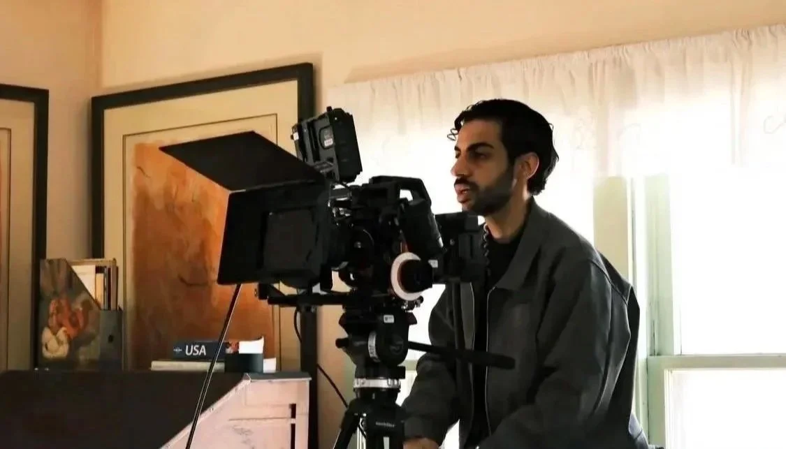 A man operating a professional video camera indoors, with artwork on the walls and a window with curtains in the background.