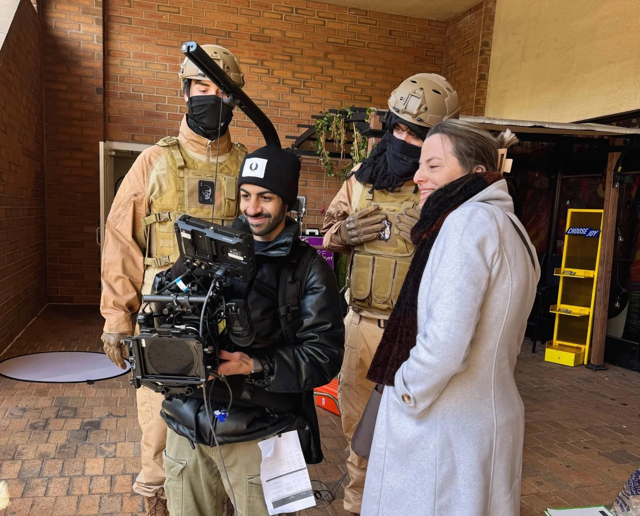 Two military personnel in tactical gear and helmets talking with a woman in a hoodie, holding a monitor with a script or checklist, in a semi-outdoor setting with brick walls and equipment.