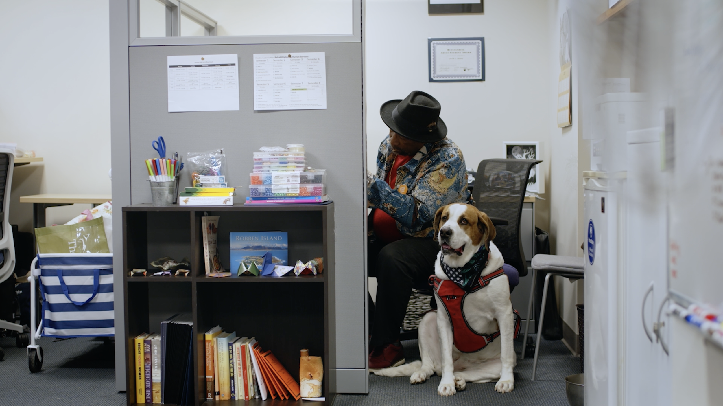 An office cubicle with a man sitting next to a dog wearing a red vest. The man is wearing a hat and patterned shirt, and appears to be looking down at something. The cubicle has various office supplies, papers, and books, as well as a certificate hanging on the wall.