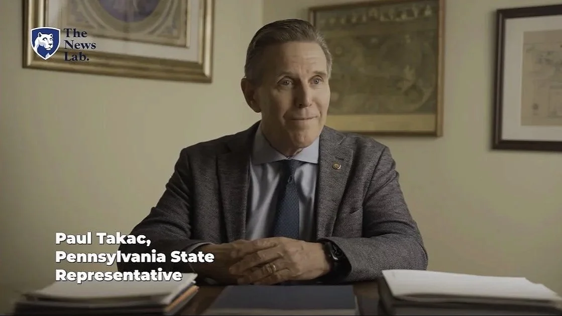 A man sitting at a desk with papers, wearing a gray blazer and blue tie, in an office with framed artwork on the wall. Text overlay identifies him as Paul Takac, Pennsylvania State Representative, with a news logo in the top left corner.