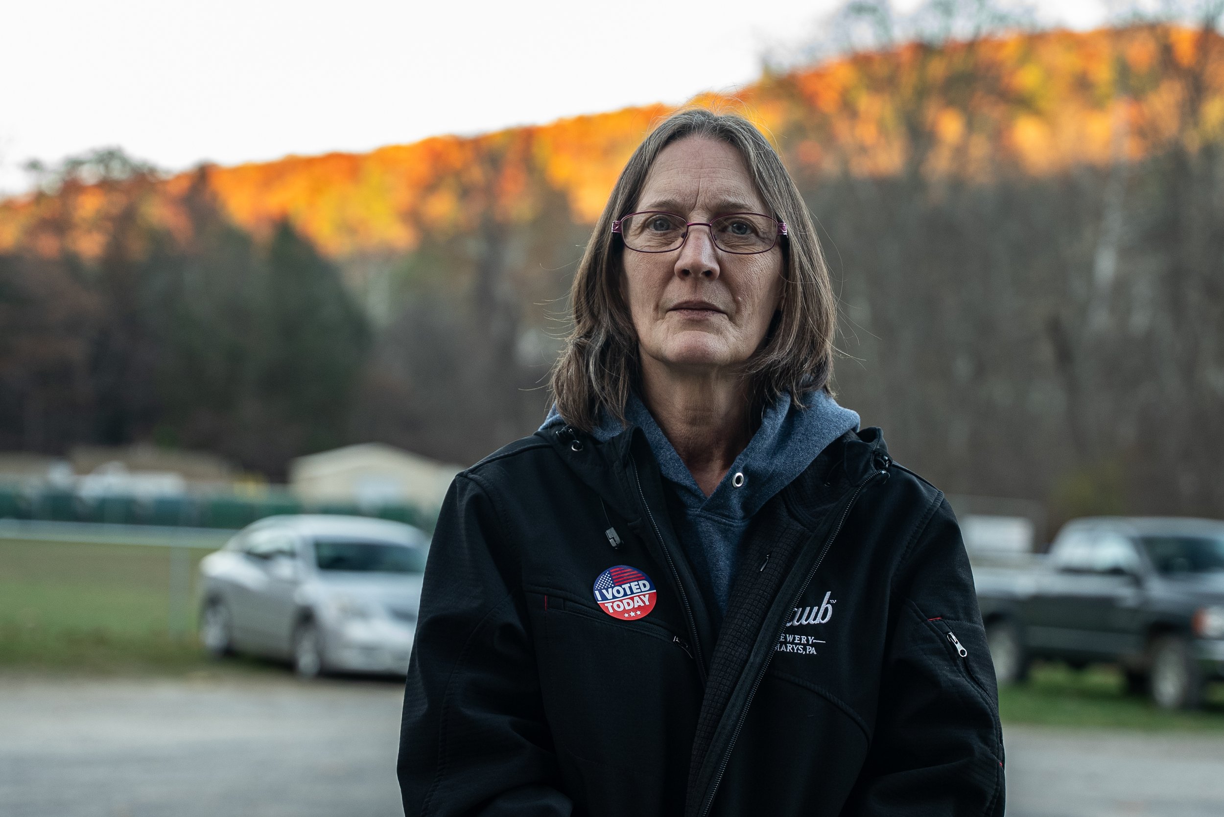 A woman with glasses and shoulder-length gray hair standing outdoors near parked cars, wearing a black jacket with an 'I Voted Today' sticker.