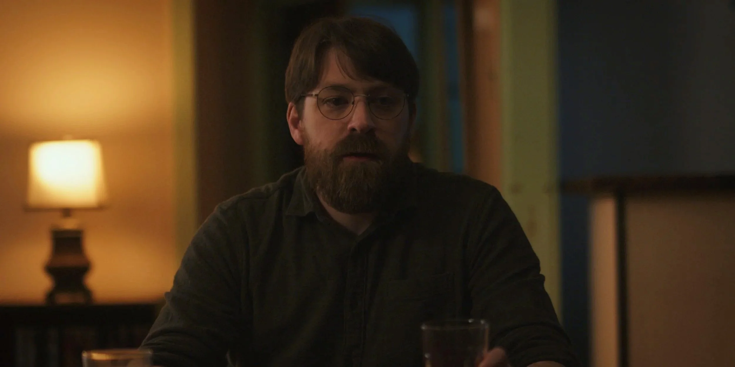 A man with dark hair, glasses, and a beard sitting at a table in a dimly lit room with warm lighting, a lamp on the side, and colorful wallpaper in the background.