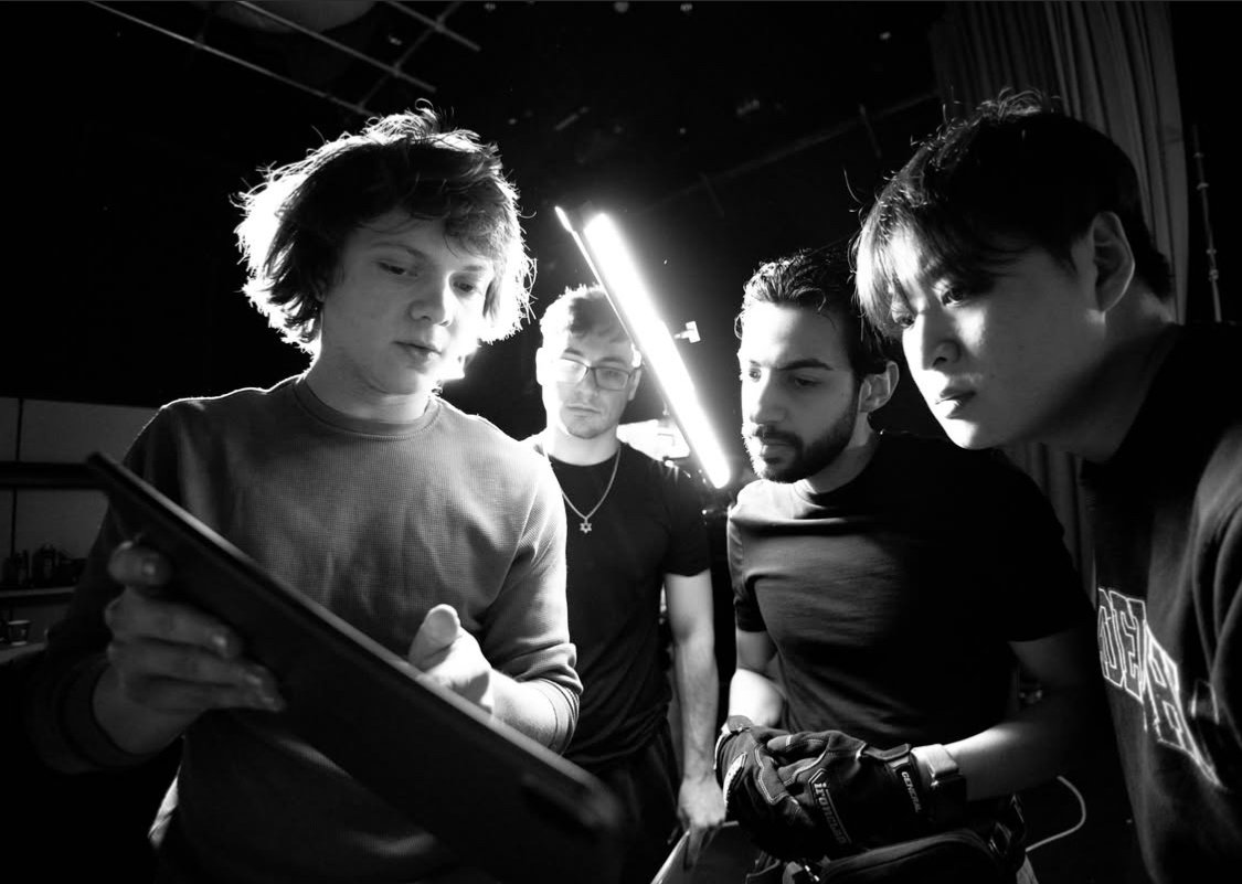 Four young men in black T-shirts are closely examining a tablet, with intense focus, in a dimly lit room with modern lighting.